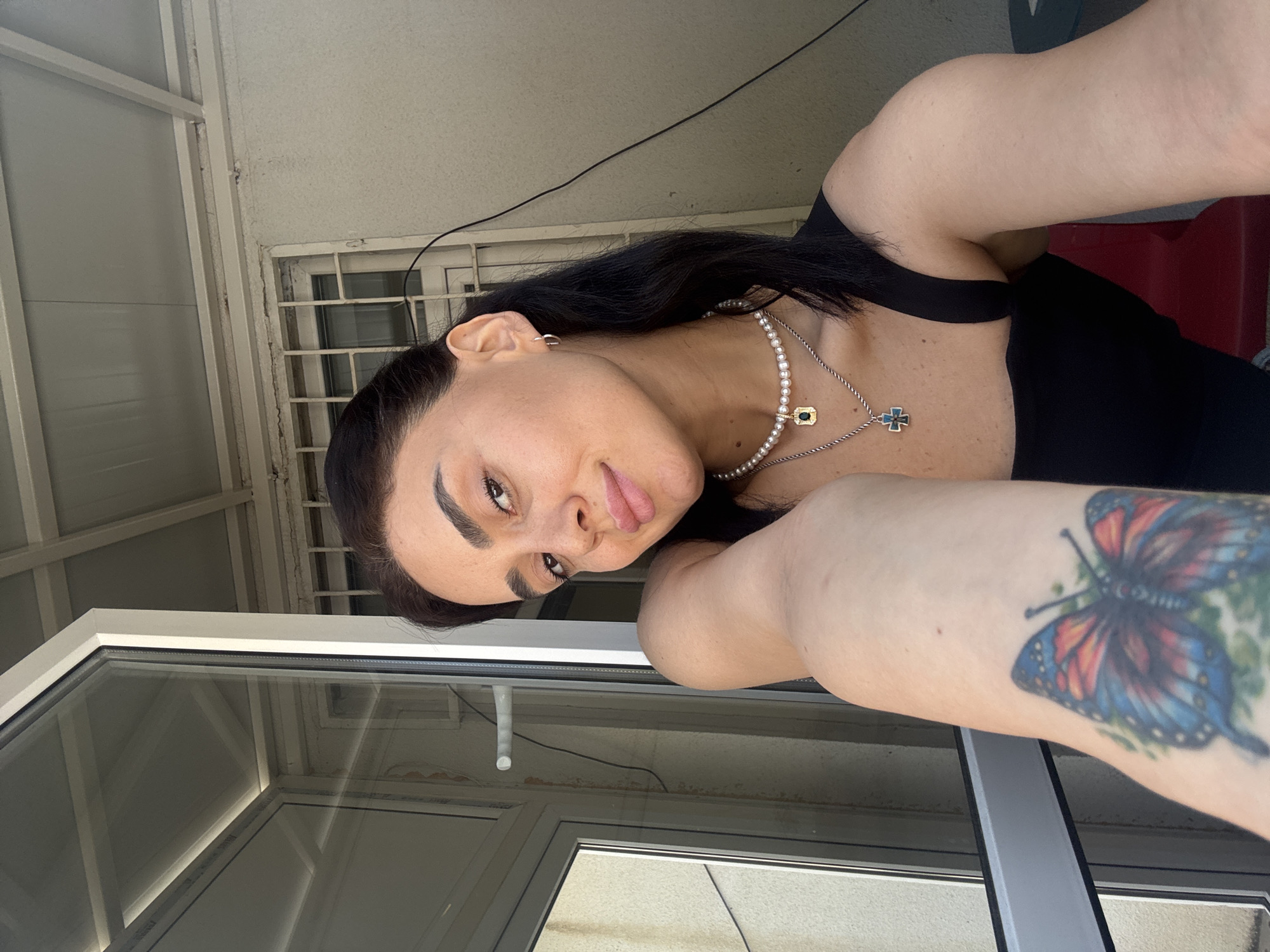 A young woman with dark hair and makeup, wearing a black tank top and necklaces, is taking a selfie. She is positioned in front of a partially open sliding glass door and a window with bars. A colorful butterfly tattoo is visible on her arm. The scene appears to be outdoors on a balcony or porch, with a glimpse of metal grating and part of a building in the background. The lighting suggests it is daytime.