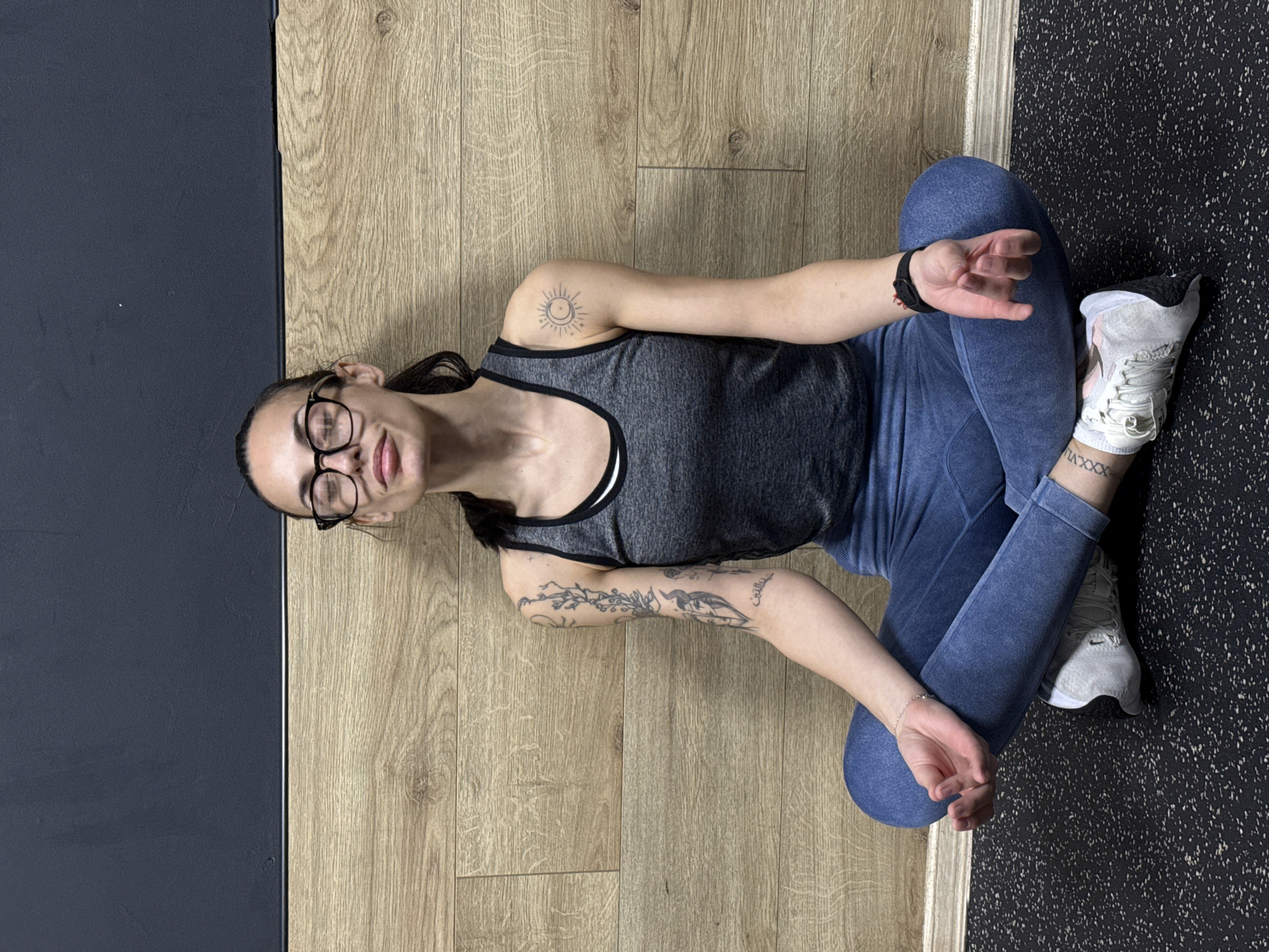 A young woman with long dark hair, glasses, and visible tattoos is sitting cross-legged on a floor in what appears to be a gym or fitness studio. She is wearing a gray tank top and blue leggings, with white running shoes visible on her feet. The floor is divided into sections: wood flooring on one side and black rubberized flooring on the other. There is a dark blue wall panel on the left side of the image. The location cannot be identified as the city of Pleven, Bulgaria, based on the visual information provided.