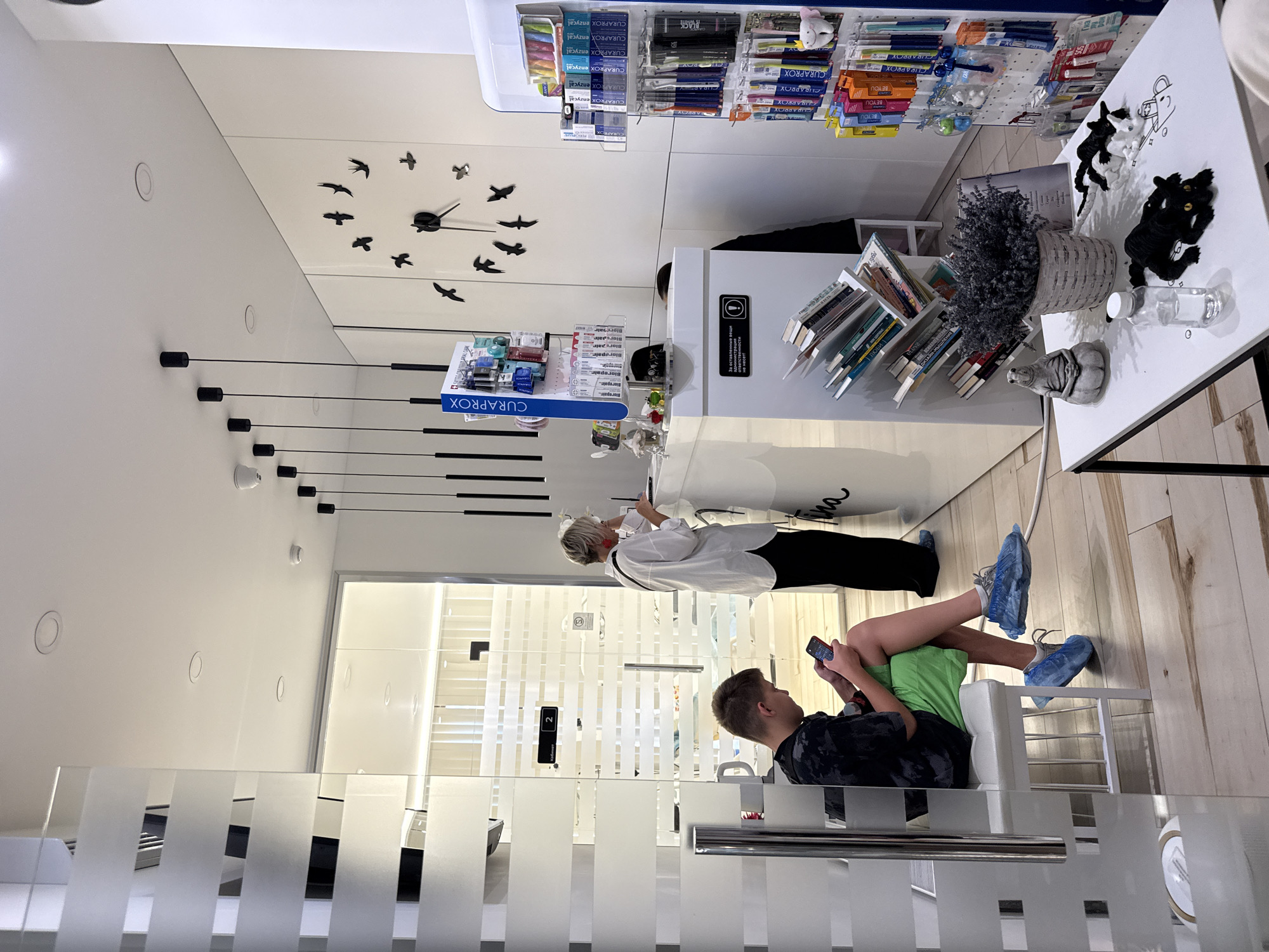 The image depicts a waiting area and reception desk inside a modern dental clinic. In the foreground, a young male is seated on a white bench, engrossed in his mobile phone. He is wearing a dark patterned t-shirt, green shorts, and blue shoe covers. To the right, a woman with blonde hair and red earrings, dressed in a white shirt and black pants, stands at the reception desk, interacting with a device or paperwork. Behind the reception desk, shelves display dental hygiene products such as toothbrushes and toothpaste. A large wall clock with a bird motif adorns the back wall. The clinic's interior features white walls and sleek furniture, with frosted glass partitions separating different areas. The floor is made of light-colored wood planks. The overall ambiance is clean and contemporary, suggesting a professional healthcare environment. The location is the reception area of a dental clinic in Мирновское сельское поселение, Ukraine.