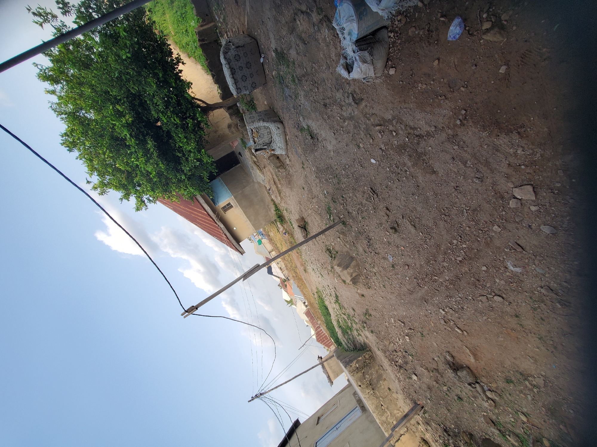 An outdoor, ground-level, wide-angle shot captures a rural village scene with a slight tilt to the left. In the foreground, a dirt and gravel path is visible, with scattered rocks and debris. To the left, a light-colored building with a blue door and window stands partially in frame. Further into the scene, a dirt embankment leads up to several structures with reddish-brown roofs. A prominent green tree with dense foliage stands to the right of the buildings, casting a shadow on the ground. Two worn, cushioned seating objects, resembling armchairs, are positioned on the ground near the tree. In the far right of the image, several sacks, some appearing to be made of burlap, are stacked. Overhead, a clear blue sky with scattered white clouds is visible, with power lines crisscrossing the frame, supported by wooden poles. The overall impression is one of a simple, somewhat unkempt, residential area.