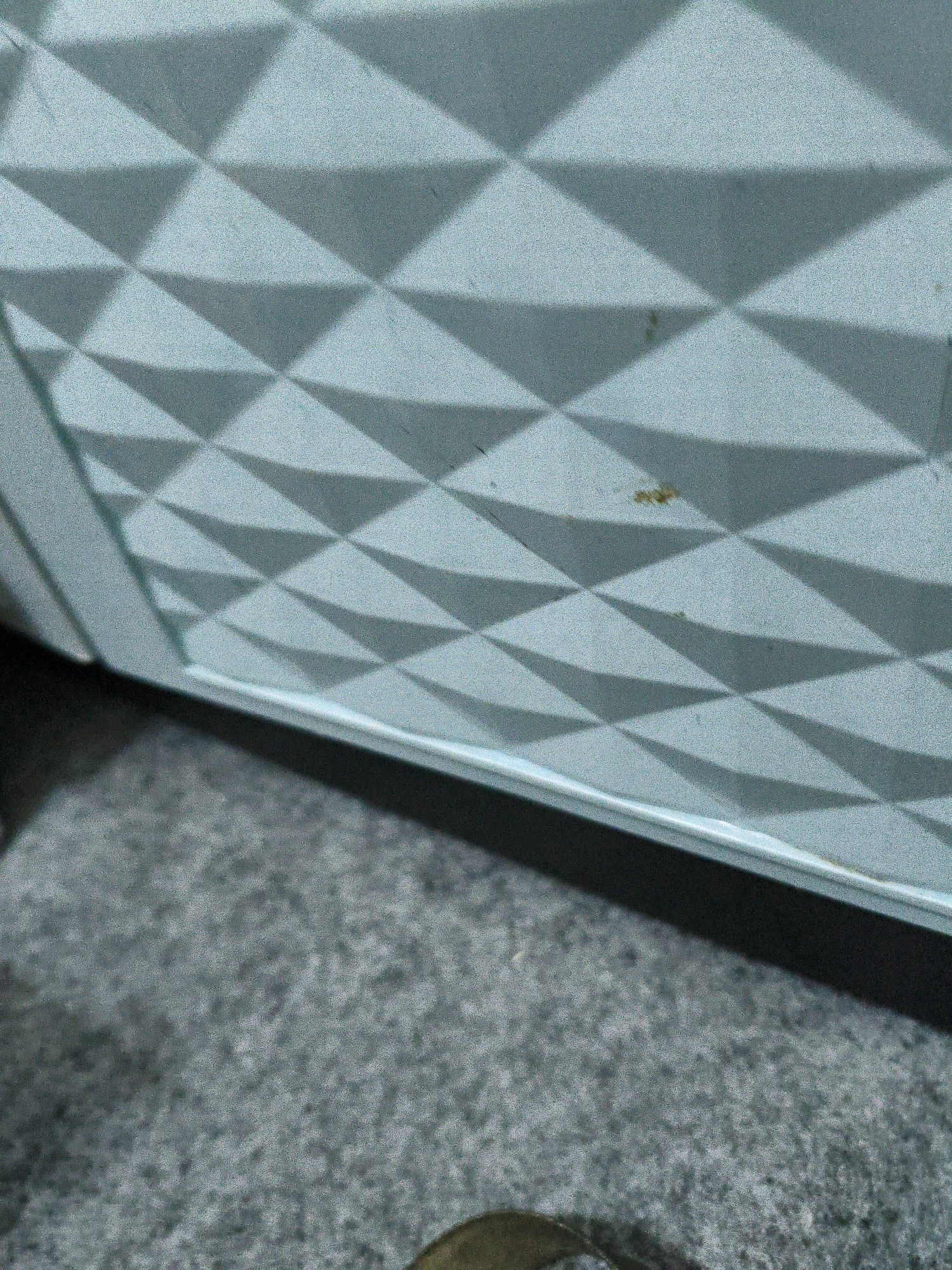 The image shows a close-up of a textured, light blue surface with a repeating diamond pattern. The surface appears to be part of an object, possibly a piece of furniture or a decorative panel, casting a shadow on the textured gray ground below. There are a few small, dark specks on the blue surface, one of which appears to be a reddish-brown stain. The bottom portion of the image shows a sliver of a metallic object, possibly a ring or a buckle, on the gray ground. The lighting suggests an indoor environment, but there is no specific indication of its location within Karangtalun, Indonesia. The scene is static and shows no activity or interaction.