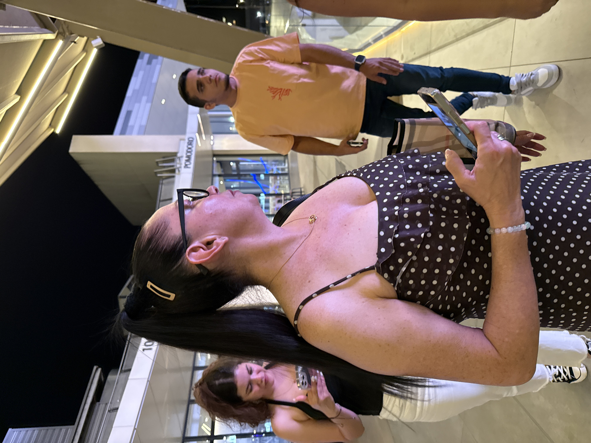 The image depicts a group of people in an urban outdoor setting at night. The primary subjects are three individuals. In the foreground, a woman with dark hair pulled back in a ponytail, wearing glasses and a brown polka dot dress, is looking upwards. Her left arm, adorned with a light green beaded bracelet, is visible, as is her right hand holding a smartphone. Behind her, another woman with reddish-brown hair is also holding a smartphone and appears to be taking a selfie. In the background, a man in an orange t-shirt and dark jeans is standing, holding his phone and looking to the side. He has a watch on his left wrist.
The setting appears to be a modern urban environment with sleek architectural elements and bright lighting. There are white beams and illuminated panels visible, suggesting a contemporary building or public space. A sign with the text "POMODORO" is visible in the background. The lighting suggests it is nighttime. The mood seems casual and relaxed, with people engaged with their phones and in conversation or observation. The overall scene suggests a social gathering or a moment captured while out and about in a city at night. The location is identified as Alcorcón, Spain.