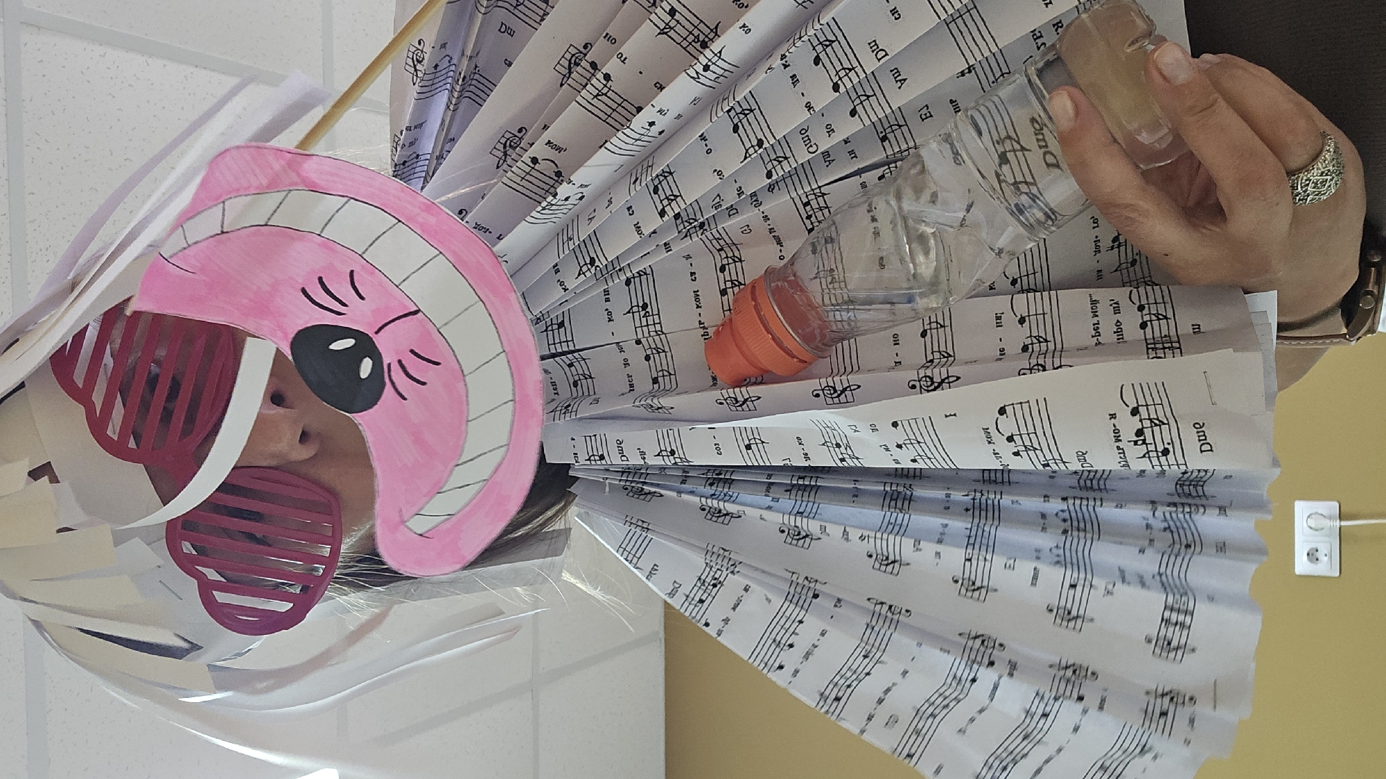 A low-angle, medium shot captures a person from the chest up, wearing a costume resembling the Cheshire Cat. The costume features a large, pink, smiling cat face with black eyes and mouth, positioned over the person's head, obscuring their face. Pink, heart-shaped sunglasses with striped lenses are worn over the person's eyes. The costume's body is made of folded sheet music, creating a voluminous, fan-like effect around the person's torso. A clear plastic bottle with an orange cap, partially filled with a light-colored liquid, is held by a person's hand, which is visible in the lower right of the frame. The hand wears a decorative ring on the ring finger. The background consists of a white drop ceiling with visible light fixtures, and a light yellow wall. An electrical outlet with a white plug is visible on the wall near the bottom of the frame. The scene appears to be indoors, possibly in a classroom or event space.