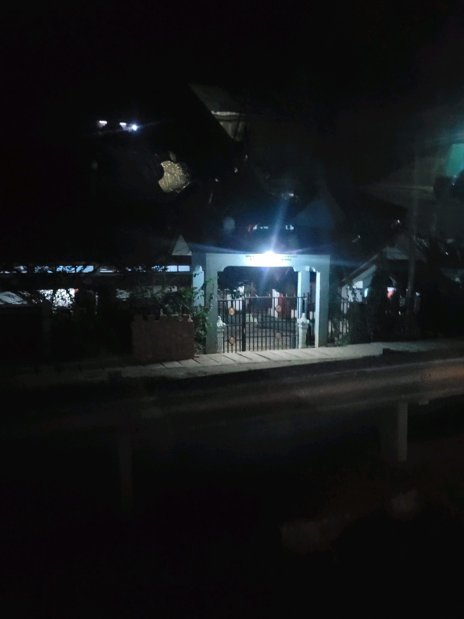The image depicts an outdoor scene at night, likely in a residential or commercial area of Kota Kuala Simpang, Indonesia. A gate with ornate metal bars is the central focus, leading into a building with a light-colored facade and a dark-colored roof. There are lights illuminating the entrance and the interior of the building, suggesting it is open or in use. Adjacent to the gate, there is a low brick wall and some foliage. The ground in front of the gate appears to be a paved walkway or sidewalk, and beyond that, a road or street is visible, with some diffused light reflecting on its surface. The overall lighting is dim, typical of nighttime conditions, with artificial lights from the building and possibly streetlights providing the primary illumination.