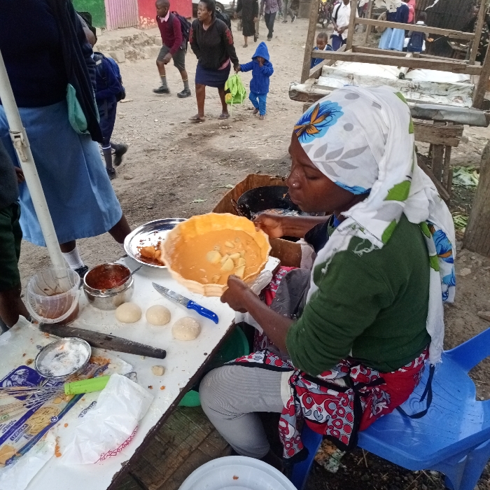 The image captures an outdoor food preparation scene in an informal settlement or bustling market area within Nairobi, Kenya. An adult female, with dark skin, is seated on a blue plastic stool. She wears a white headscarf with blue and green floral patterns, a dark green long-sleeved shirt, light-colored pants, and a red-and-white patterned apron. Her hands are actively engaged in preparing food, holding a large, shallow, round metal pan containing an orange-brown liquid with lighter-colored solid chunks, surrounded by a ruffled, fried pastry. On a white surface in front of her are multiple small, round dough balls, a silver knife with a blue handle, a small metal bowl with a reddish sauce, a clear plastic container with a brown liquid, a metal plate with remnants of fried food, a wooden rolling pin, and various plastic packaging materials. A dark cooking pot is partially visible to her right. In the background, on an unpaved dirt path, several individuals are walking. These include school children in dark blue and maroon uniforms, an adult woman holding hands with a small child dressed in a blue hooded jacket, and other pedestrians. The background also features simple structures with corrugated iron or painted walls in various colors, indicative of an informal urban environment.