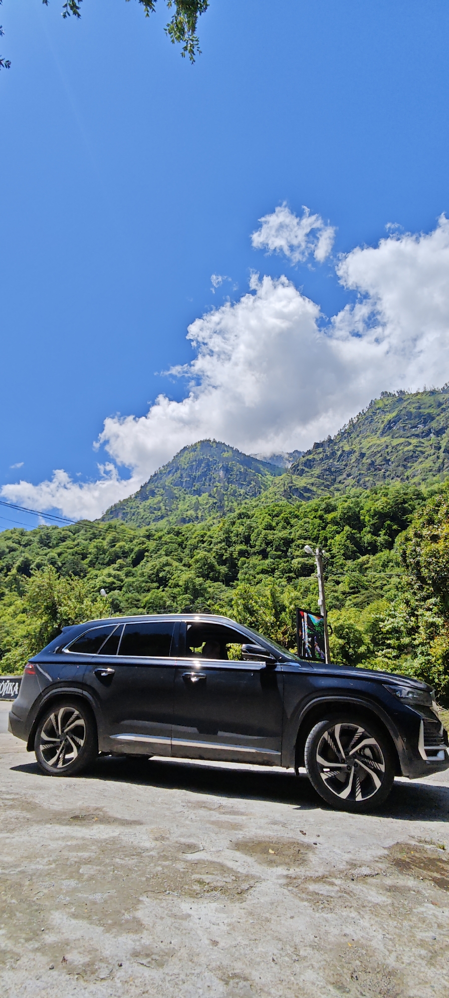 A dark-colored SUV, likely black or dark grey, is parked on a light-colored paved surface by the side of a road, set against a stunning backdrop of lush green, forested mountains. The car features tinted windows, silver trim along its windows and lower body, and distinctive alloy wheels with a swirling, multi-spoke design. A person's silhouette is faintly visible through the driver's side window.
The mountains are densely covered in vibrant green trees, appearing steep and rugged, with some peaks touching the bright blue sky. Numerous white, fluffy clouds are scattered across the sky, particularly concentrated around the mountain peaks on the right. Bright sunlight illuminates the scene, suggesting a clear day, likely morning or early afternoon, with a subtle sun flare visible in the upper left corner of the sky. A few green tree branches hang into the frame from the very top left.
Near the front of the car, a tall street light pole is visible, and beneath it, an electronic sign or screen displays a colorful, somewhat glitched pattern. The paved ground beneath the car shows some wear and patches of dampness. On the far left, partially obscured by the car's front door, a white sign with Cyrillic letters is visible, showing "ЙКА," which could be part of a word like "СТОЙКА" (meaning "stand" or "counter"). This scenic moment is captured in a mountainous region, specifically Bzypta, Abkhazia, highlighting a travel stop amidst natural beauty.