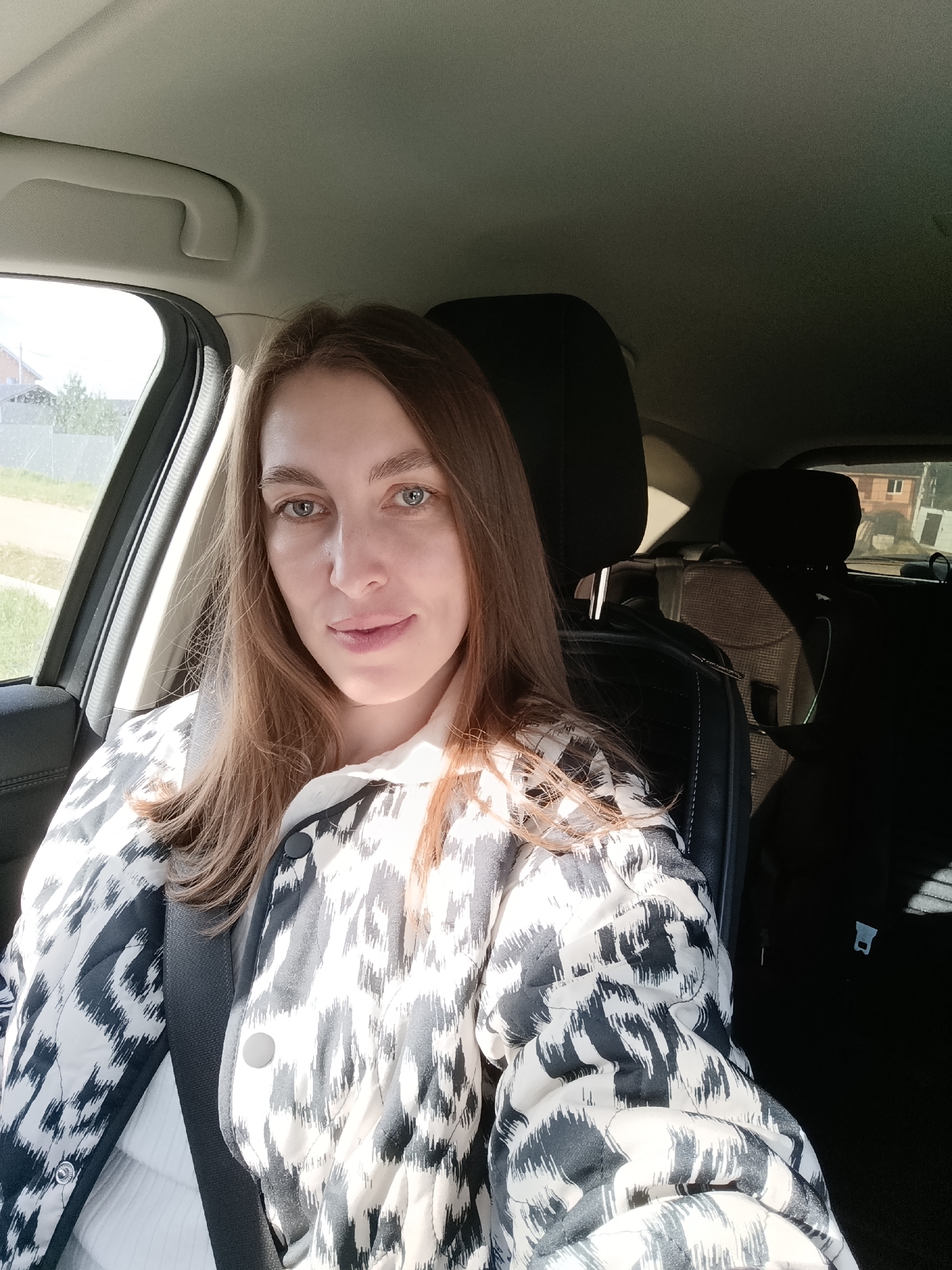 A woman is sitting in the driver's seat of a car, wearing a black and white patterned jacket over a white top, secured by a seatbelt. Outside the window, the landscape suggests a suburban area, with a fence, trees, and the corner of a building visible. The interior of the car is dark, except for the front seat area illuminated by sunlight. The location appears to be Chaikovsky, Russia, based on geographical context and the design of the car interior.