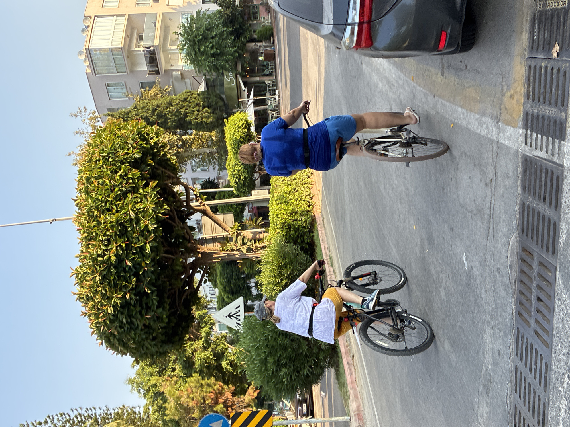 In Alanya, Turkey, two individuals are seen cycling on a paved road. The person in the foreground, facing away from the camera, is wearing a blue t-shirt and blue shorts, riding a bicycle. To their left, another person, also facing away from the camera, is wearing a white t-shirt, yellow shorts, and a camouflage cap, riding a bicycle. Both individuals are on the right side of the road. The environment includes a street lined with trees and bushes, a pedestrian crossing sign, buildings in the background, and a parked car on the right side of the road. The sky is clear and blue.