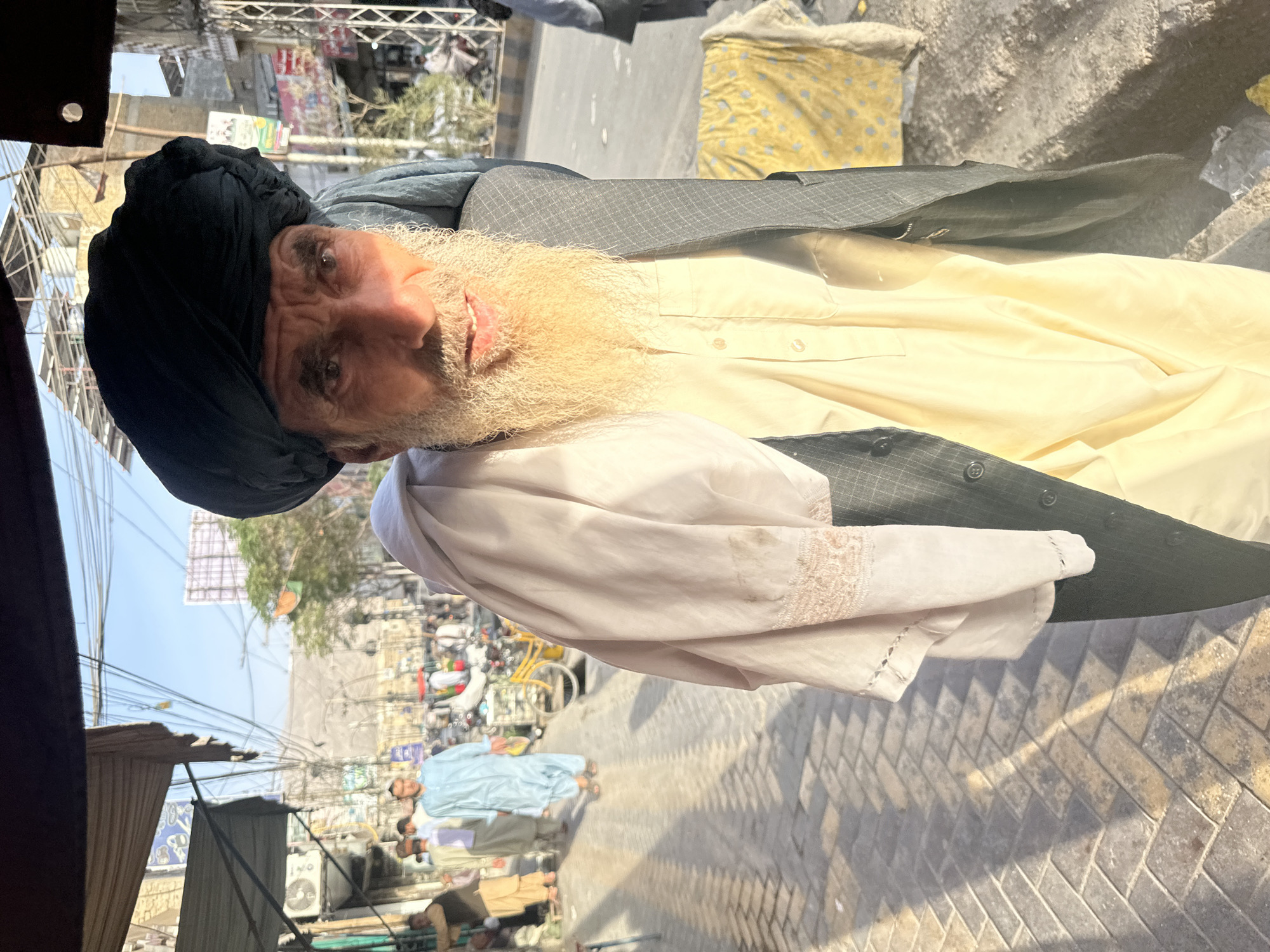 An elderly man with a long white beard and a dark turban stands on a paved sidewalk in Quetta City Tehsil, Pakistan. He is wearing a light yellow shirt and a dark, pinstriped jacket, draped with a light-colored fabric. Sunlight casts long shadows across the sidewalk. In the background, a bustling street scene unfolds, with other pedestrians, market stalls, a bicycle, and various structures. Overhead, a dense network of wires crisscrosses the sky, obscuring parts of the buildings and the sky above. The overall atmosphere suggests a vibrant, sun-drenched urban environment.