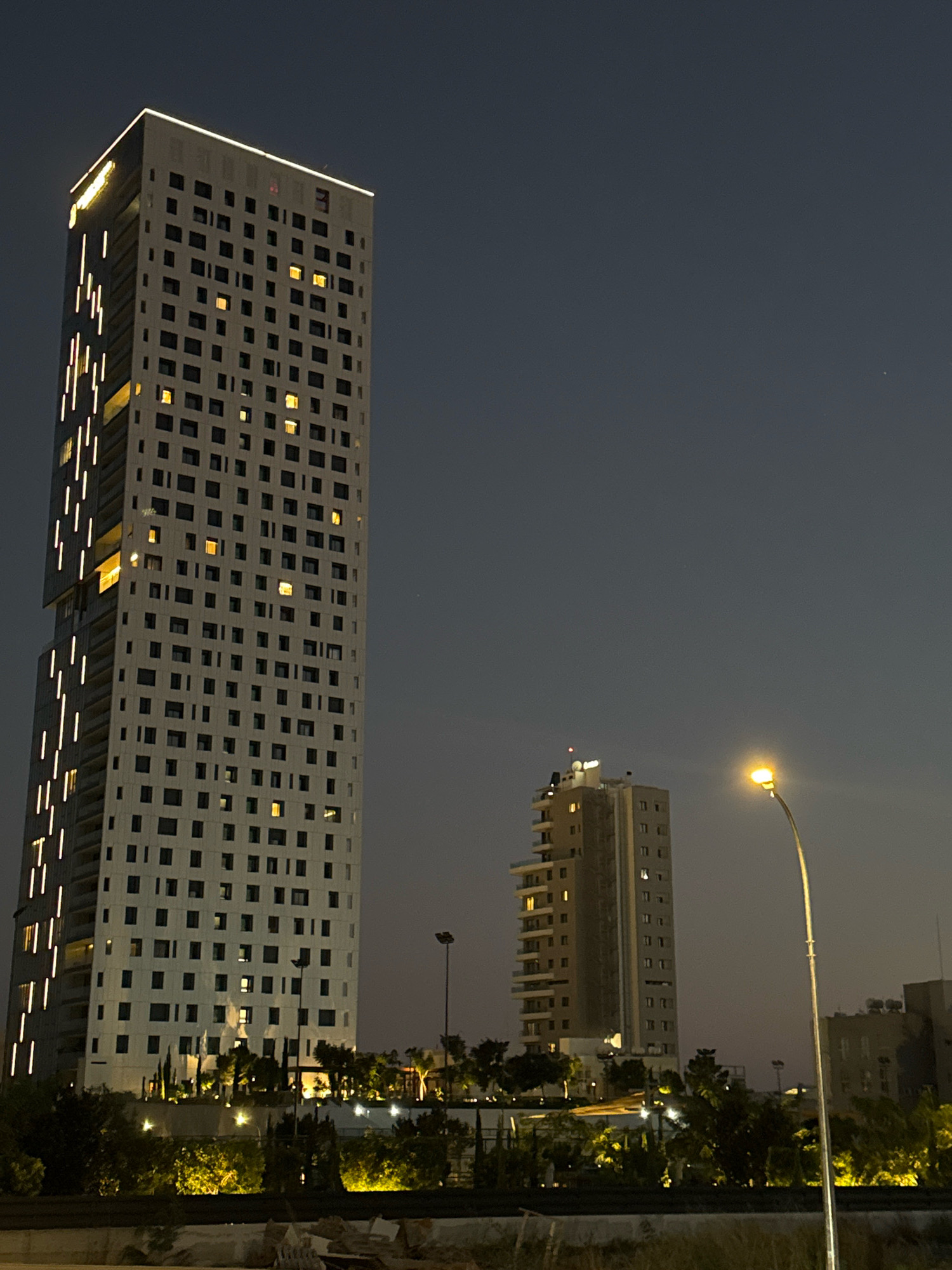 In the Mouttagiaka Community, Cyprus, a night scene is depicted. The foreground features a dark, textured surface, possibly a road or embankment, with some debris visible. Behind this, a landscaped area with illuminated trees and foliage creates a warm glow. Two distinct buildings dominate the mid-ground and background.
The primary structure on the left is a tall, modern, rectangular skyscraper with a white facade. It is characterized by a grid of dark square windows and vertical strips of bright white lights running along its left side. The top of this building is outlined with a line of white lights, and a yellow illuminated sign is visible near the top left corner.
To the right of the skyscraper, a slightly shorter, lighter-colored rectangular building stands. This building also has numerous dark windows, some of which are illuminated with a warm, yellow light.
A streetlamp with a bright yellow light source is positioned in the mid-right of the image, casting its illumination outwards. In the far background, other buildings are partially visible, suggesting an urban or semi-urban setting. The sky is a dark, gradient twilight hue, with a subtle haziness. There are no people or animals visible in the frame.