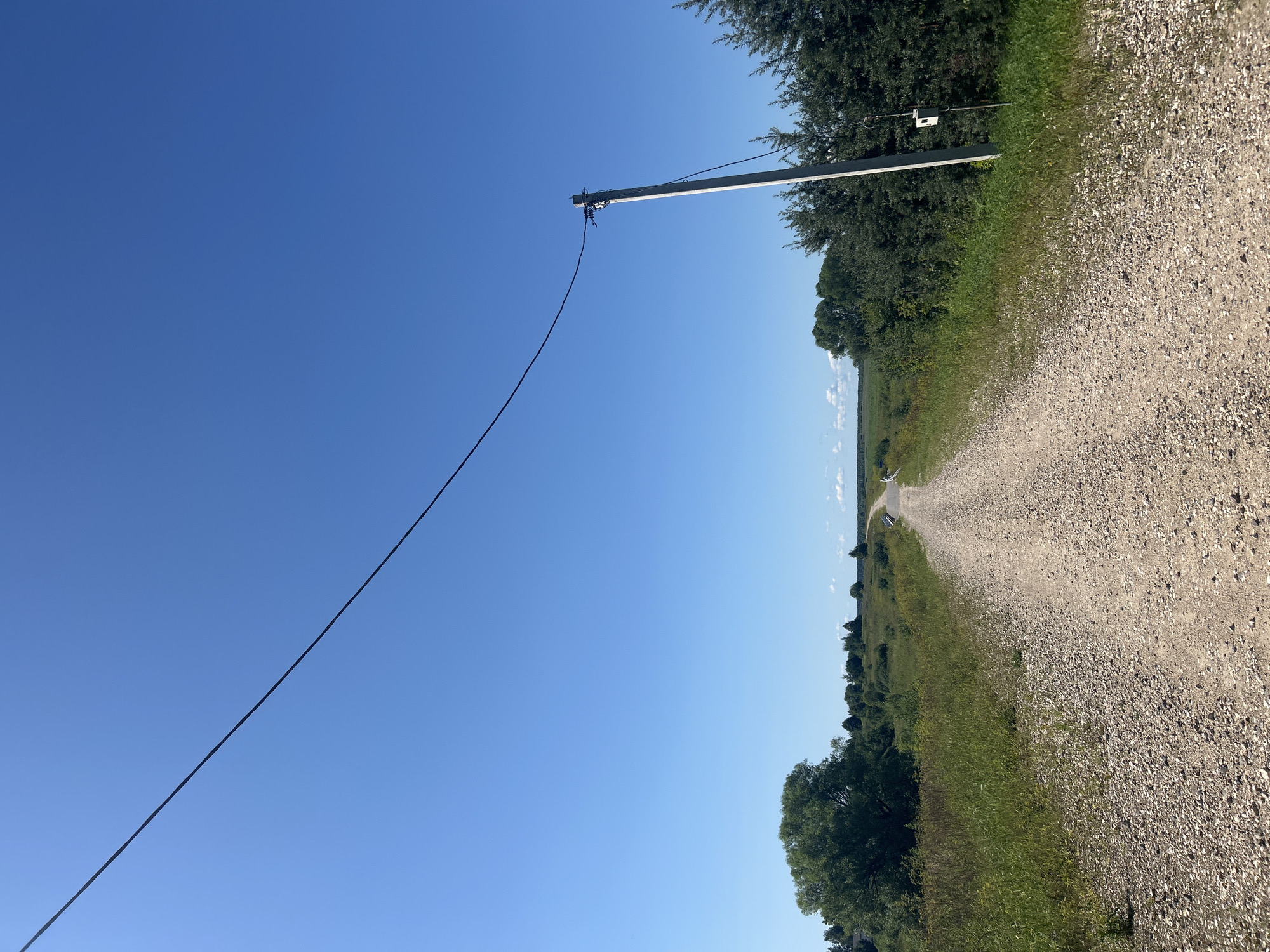 The image depicts a rural, gravel road extending into the distance under a clear blue sky. The road is flanked by green grass and various trees, with a power line stretching diagonally across the upper portion of the frame. A utility pole with some equipment attached stands to the right of the road. The landscape appears to be a natural, undeveloped area, possibly on the outskirts of the village of Kalitsino, Russia. There are no people or animals visible in the scene.
