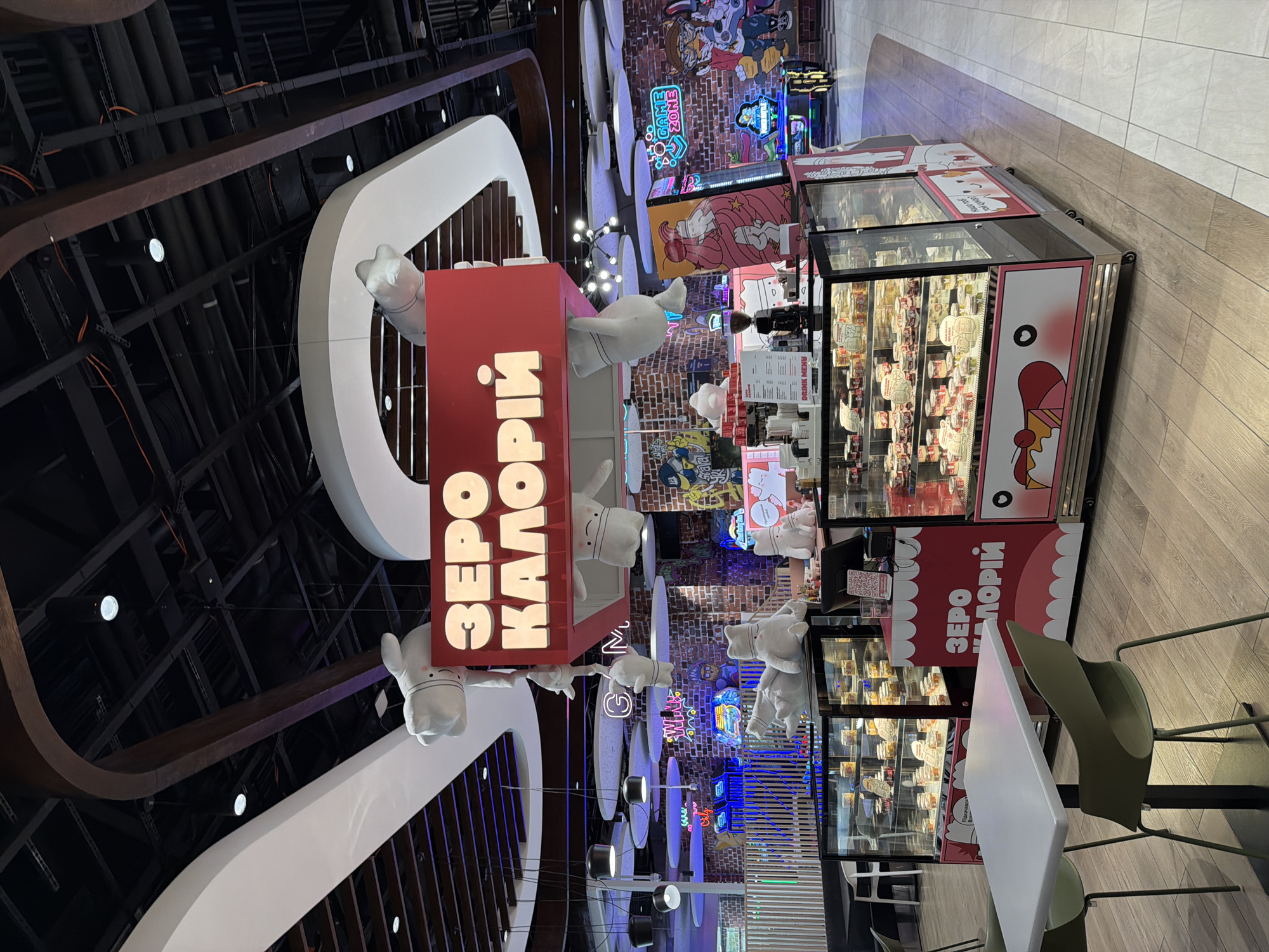 The image shows a food stall with the name "ZERO CALORII" in large, illuminated letters. The stall appears to be located in a shopping mall or entertainment center, indicated by the brick wall in the background with neon signs and arcade games. The stall itself features a refrigerated display case filled with various desserts in individual containers. There are also plush cartoon characters hanging from the ceiling and placed around the stall. In the foreground, there is a white table with two green chairs. The overall aesthetic suggests a playful and modern environment, likely targeting a younger demographic or those seeking novelty. The specific location is not identifiable from the image.