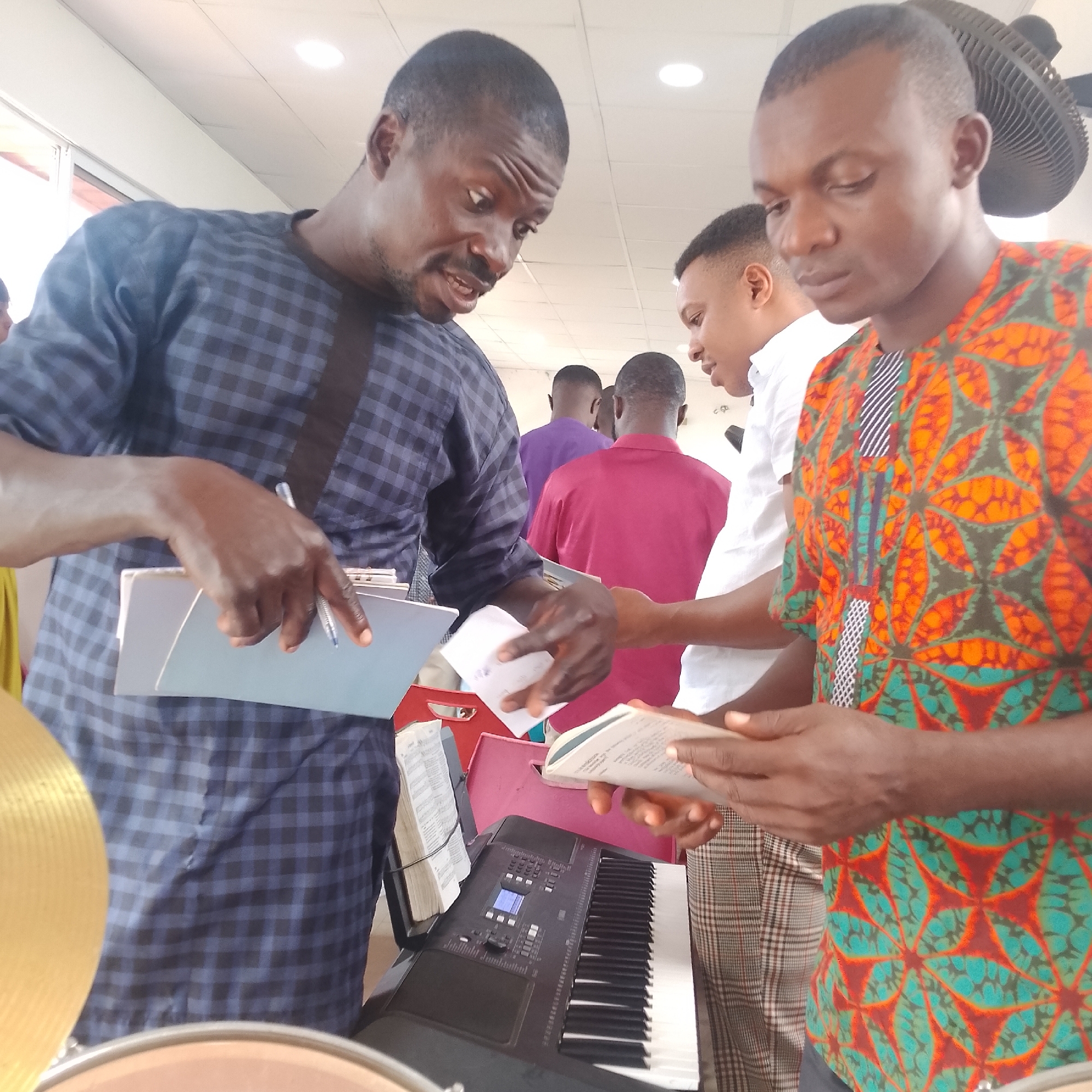 The image depicts an indoor scene at the Iranla, Nigeria location. Three men are the central figures. The man on the left, wearing a blue and black checkered shirt, is looking down and speaking while holding a pen over an open folder or binder. To his right, a man in a colorful shirt with an orange, green, and teal pattern is looking down at a book he is holding. Between these two men, another man in a white shirt is partially visible. In the background, two other men are visible from behind, one in a purple shirt and another in a maroon shirt. In the foreground, a black electronic keyboard is positioned, with an open book on its stand to the left of the keyboard. Part of a drum cymbal is visible in the lower left corner. The environment appears to be a room with a tiled ceiling and fluorescent lighting.