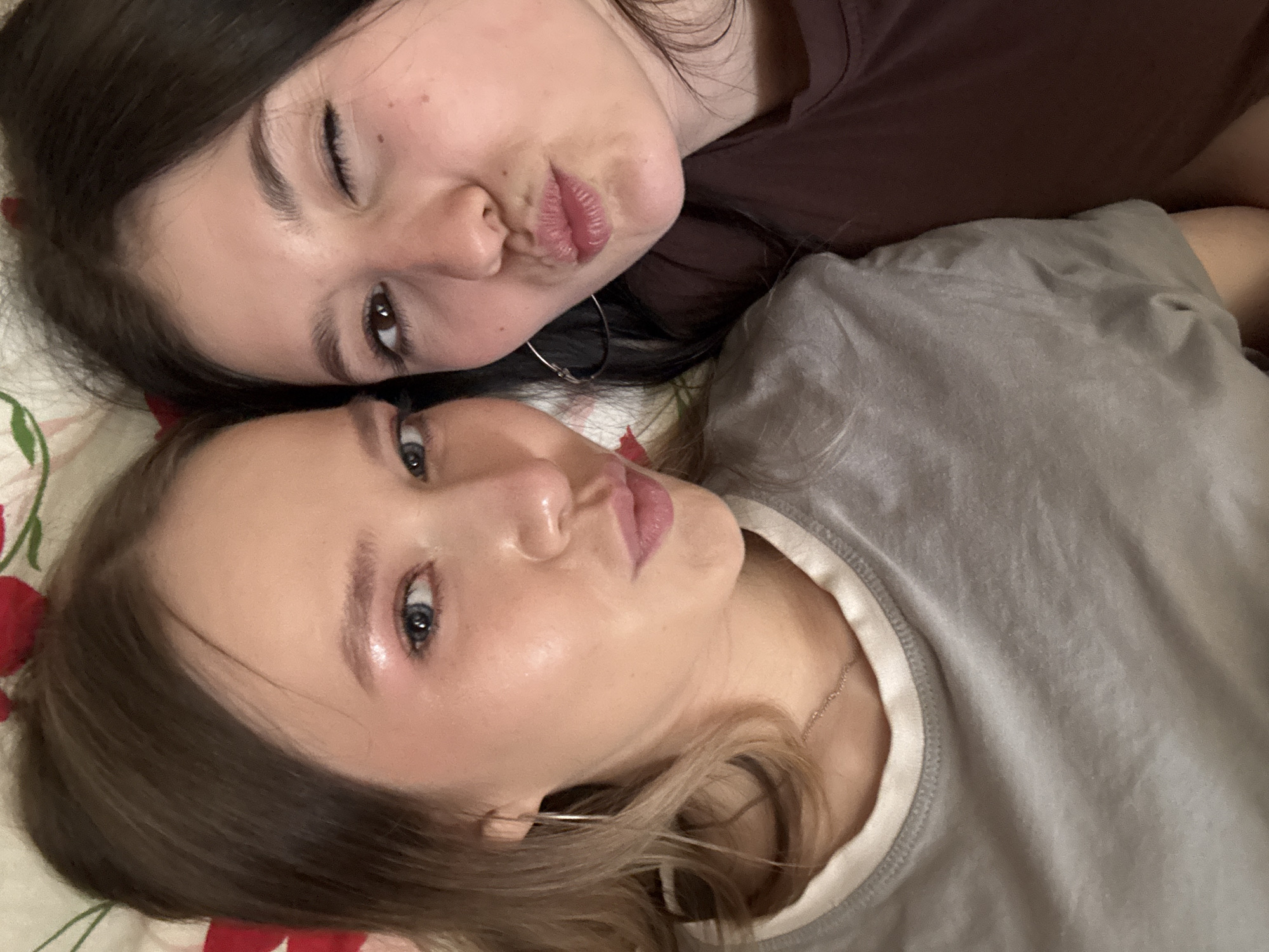 Two young women are lying down, posing for a selfie. The woman on top has dark hair and is winking, puckering her lips as if for a kiss. She wears a dark shirt and a silver hoop earring. The woman below has light brown hair and light eyes, and is also puckering her lips. She is wearing a light grey shirt with a white trim and a thin necklace. They appear to be lying on a bed with a floral patterned sheet. There is no evidence to confirm the location is Фоки, Russia.