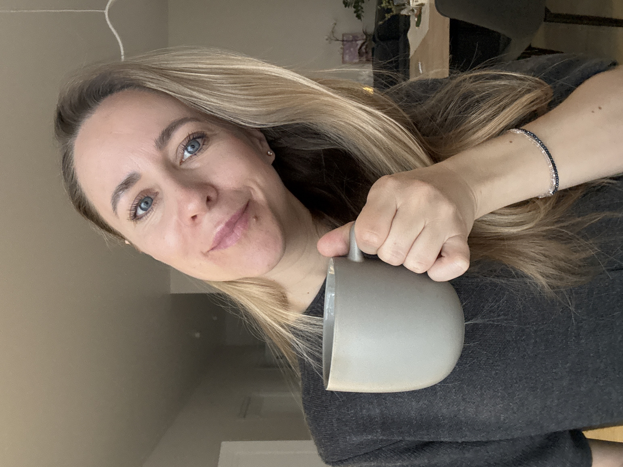 A woman with blonde hair and blue eyes is holding a gray mug. She's wearing a dark gray sweater and a beaded bracelet. The background appears to be an indoor setting, possibly a kitchen or dining area. The location is not possible to determine from the information in this image.
