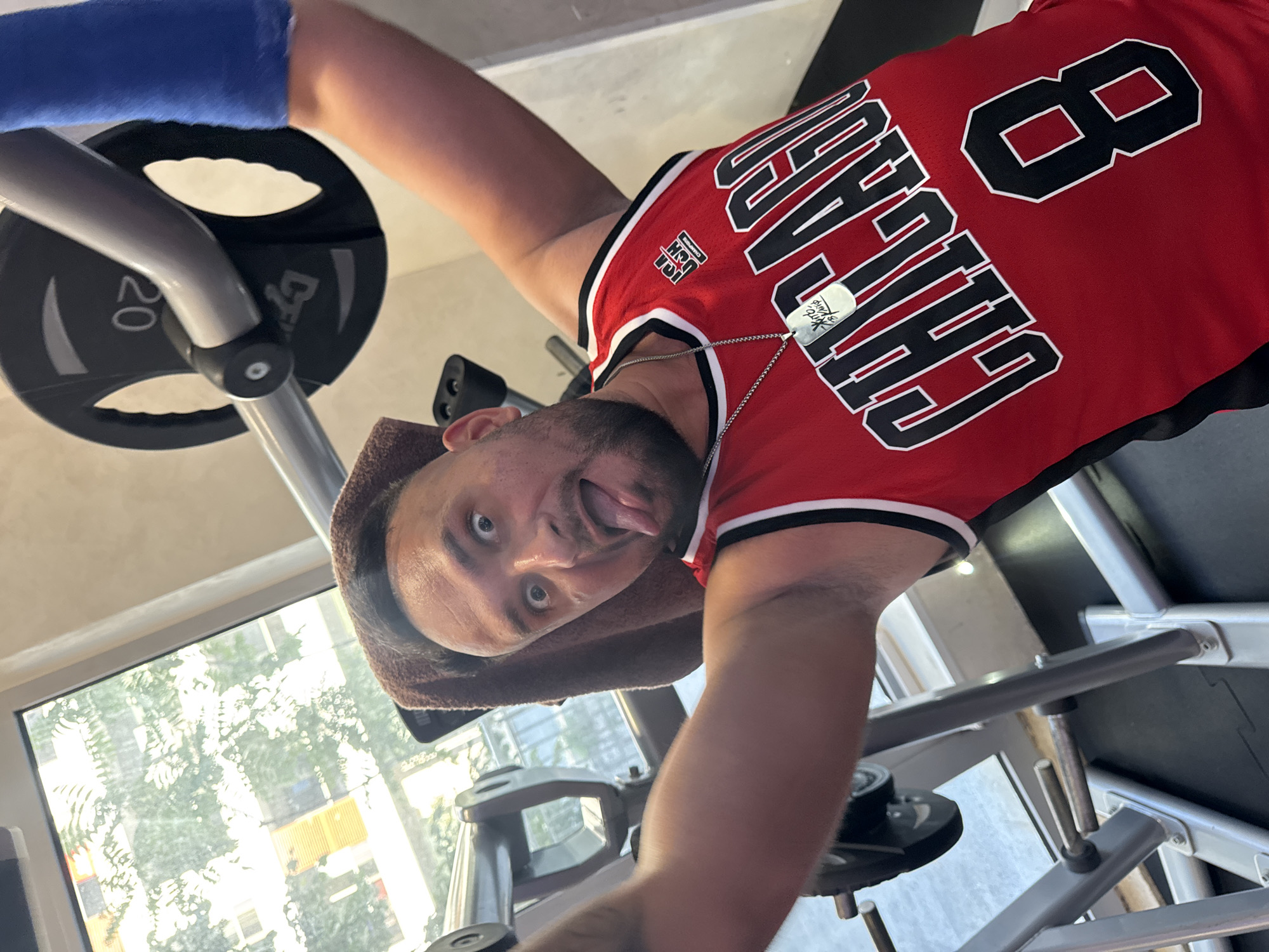 The image shows a man in a gym in Budva, Montenegro, taking a selfie while lifting weights. He is wearing a red Chicago Bulls basketball jersey with the number 8 on it, and a silver dog tag necklace. He has a towel draped over his head and is sticking his tongue out at the camera. A weight plate with the number 20 is visible on the barbell he is lifting. The background shows a gym interior with gym equipment and a window looking out onto trees and buildings.