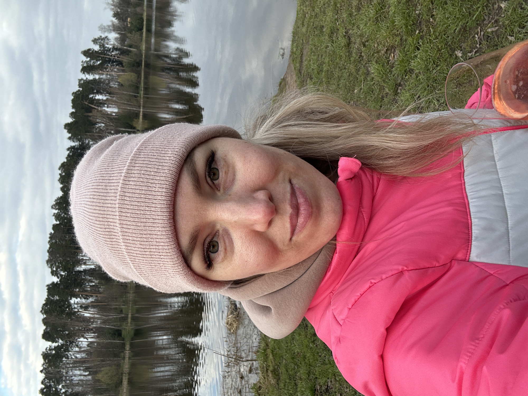 A woman in a pink beanie and matching pink jacket stands near a lake in the city of слобода Старая, Russia. The lake reflects trees and a cloudy sky. She is wearing a beige scarf and has blonde hair. A pink glass is visible to the right of the frame. Grass grows along the lake shore.