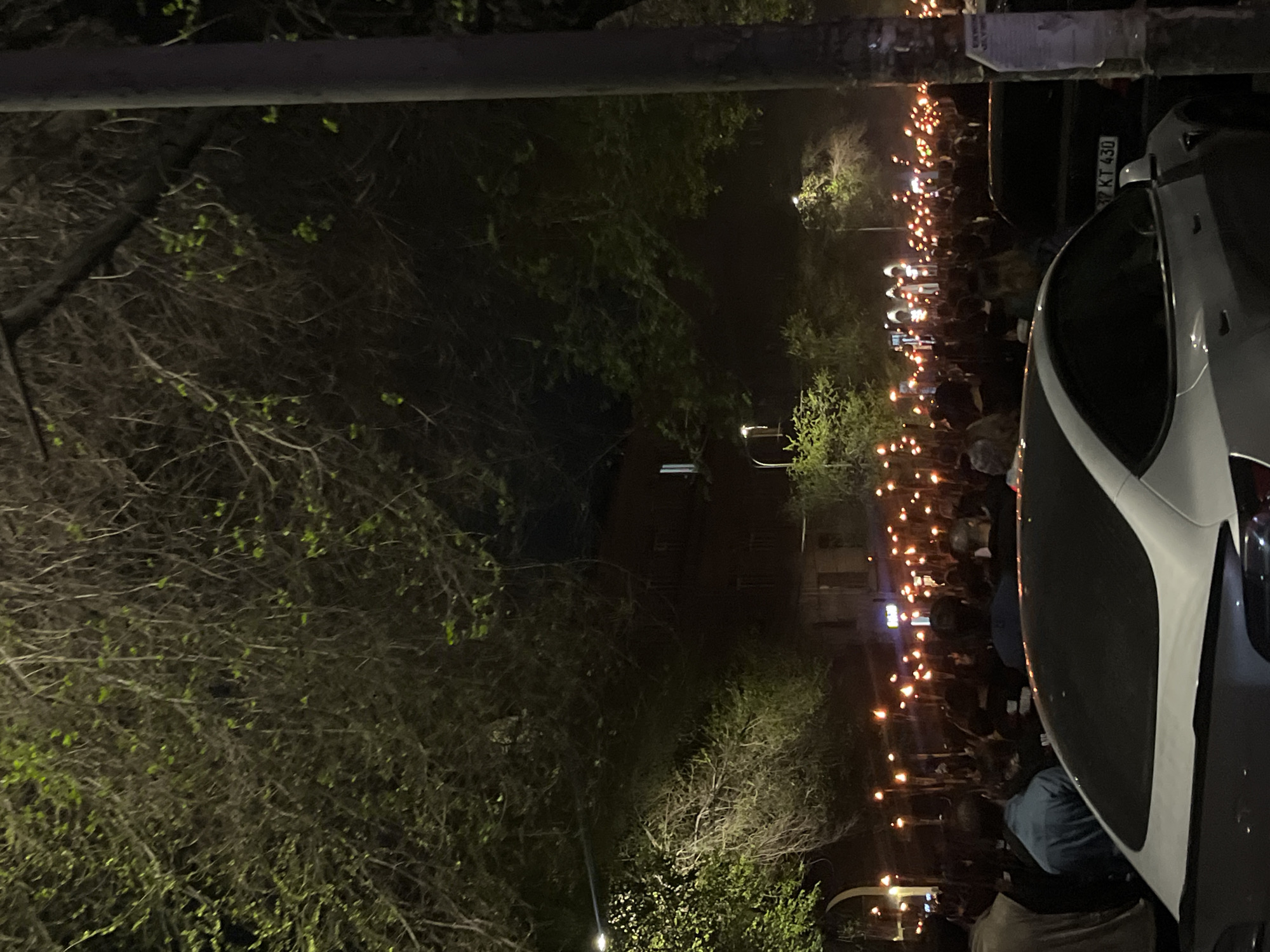 An outdoor night scene depicts a large crowd of people gathered, presumably for a procession or vigil. The image is taken from an elevated perspective, looking down and across a street or public area.
In the right half of the image, numerous individuals are visible, extending into the background. Most are holding small, glowing lights, appearing to be candles or torches, creating a warm, scattered illumination against the darkness. The figures are largely obscured by the low light and distance, but their collective presence is evident.
To the immediate right, partially visible in the foreground, is the front-right section of a white car with dark accents. Behind it, further into the scene, is a darker vehicle with a license plate clearly reading "37 KT 430." The "37" prefix on the license plate indicates that the vehicle is registered in Yerevan, Armenia, confirming the given location context.
The left and foreground portions of the image are dominated by trees and bushes, some with sparse branches and others showing signs of fresh, light green foliage. A dark, horizontal structure, possibly a pole or beam, runs across the upper part of the frame. In the distant background, dimly lit buildings can be discerned above the heads of the crowd. The overall setting is an urban environment under the cover of night, with artificial lights from the candles and some distant streetlights providing the primary illumination. The atmosphere appears to be solemn or commemorative due to the nature of the gathering and the use of candles.
