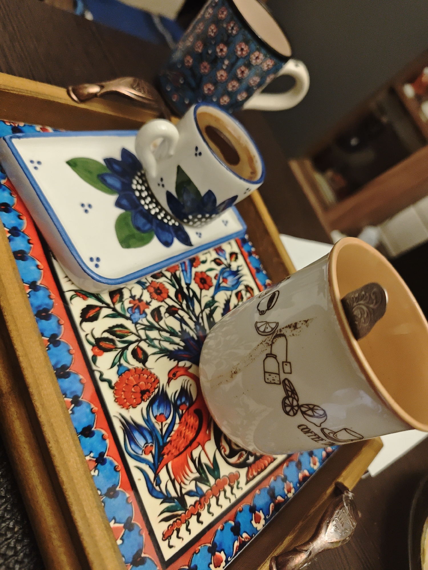 This close-up shot presents an artfully arranged coffee setup on a wooden tray. The central focus is a small ceramic coffee cup, holding dark liquid, resting on a square saucer. Both the cup and saucer feature a vibrant blue and green floral pattern against a white background. Beneath this, a larger, highly decorative ceramic tile serves as a base, exhibiting intricate patterns of red and blue flowers, green leaves, and a stylized red bird with blue accents. This tile is bordered by a bright red band with repeating blue semicircular motifs.
To the right, a larger, light-colored ceramic mug lies on its side. It is decorated with minimalist line drawings of lemons, teapots, and other beverage-related items, and the word "COFFEE" is partially visible. An ornate, dark-colored spoon is placed inside this mug. The wooden tray itself has metallic, coppery handles. In the blurred background, another blue ceramic mug with floral patterns is discernible, along with indistinct furniture. The distinct folk art style of the ceramic patterns suggests a cultural influence, possibly from the Middle East or Eastern Europe, fitting within the broader Russian context of Свистуха (кузяевский С/о). The scene is set indoors, depicting a moment of rest or an inviting beverage presentation. There are no people present, and the time of day is not evident.