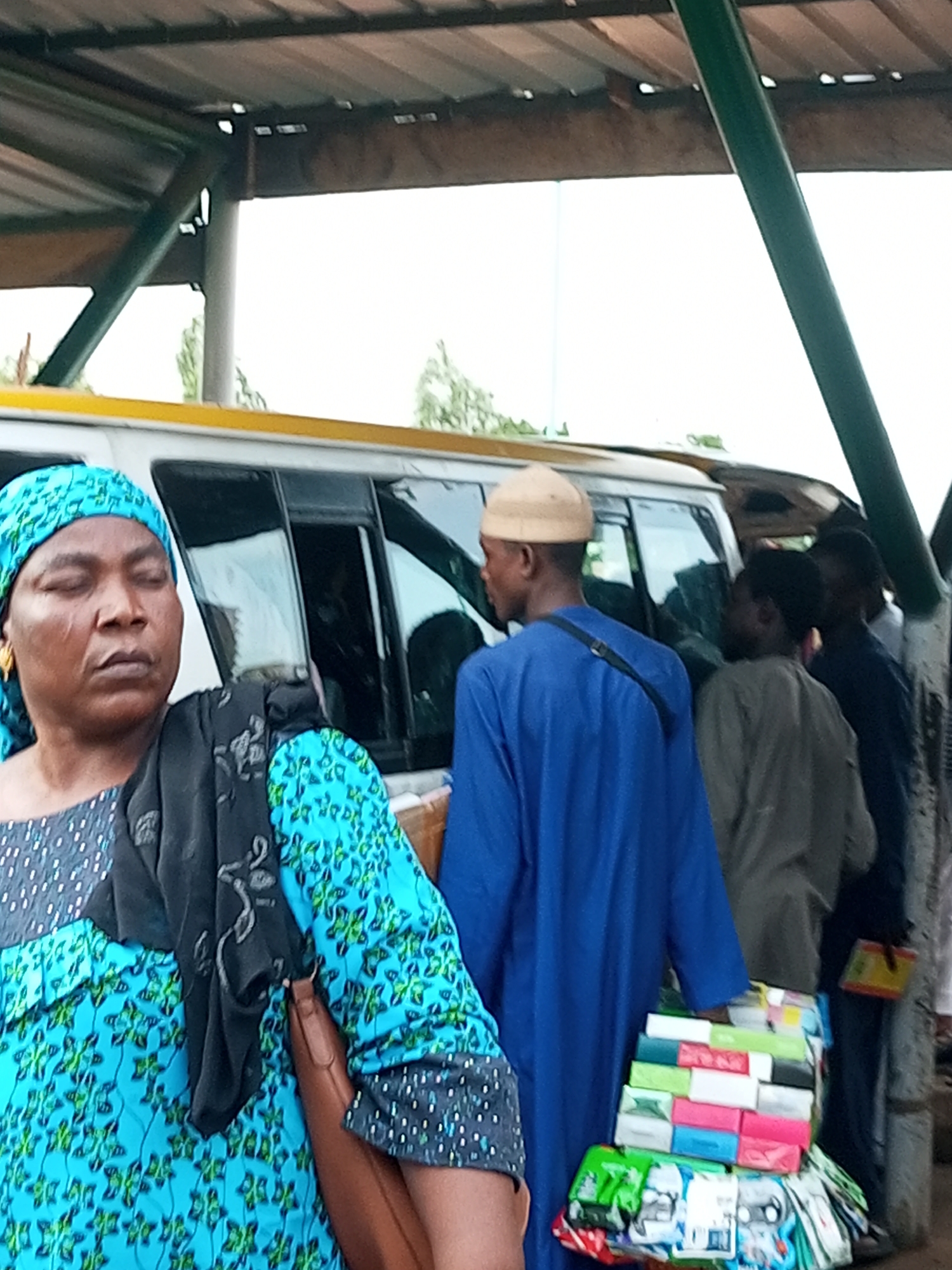 In Gombe City, Nigeria, an outdoor scene unfolds under a shaded structure, possibly a bus stop or market stall. A woman, wearing a vibrant blue and green patterned dress and a matching headscarf, stands with her eyes closed, her face conveying a sense of weariness or contemplation. She is adorned with a gold earring. Beside her, a man in a royal blue tunic and a light beige cap is partially visible, with a strap from a bag crossing his shoulder. Further back, several other individuals are present. One man wears a light olive-colored garment, while another in the background is dressed in a dark blue shirt. To the right, a collection of colorful items, likely products for sale, are stacked and displayed. These include items in bright pink, blue, white, and green packaging, some of which appear to be small boxes or packets. The backdrop is dominated by the white exterior of a vehicle, possibly a bus, with dark-tinted windows. The overall environment suggests a public gathering space where commerce and transit intersect.