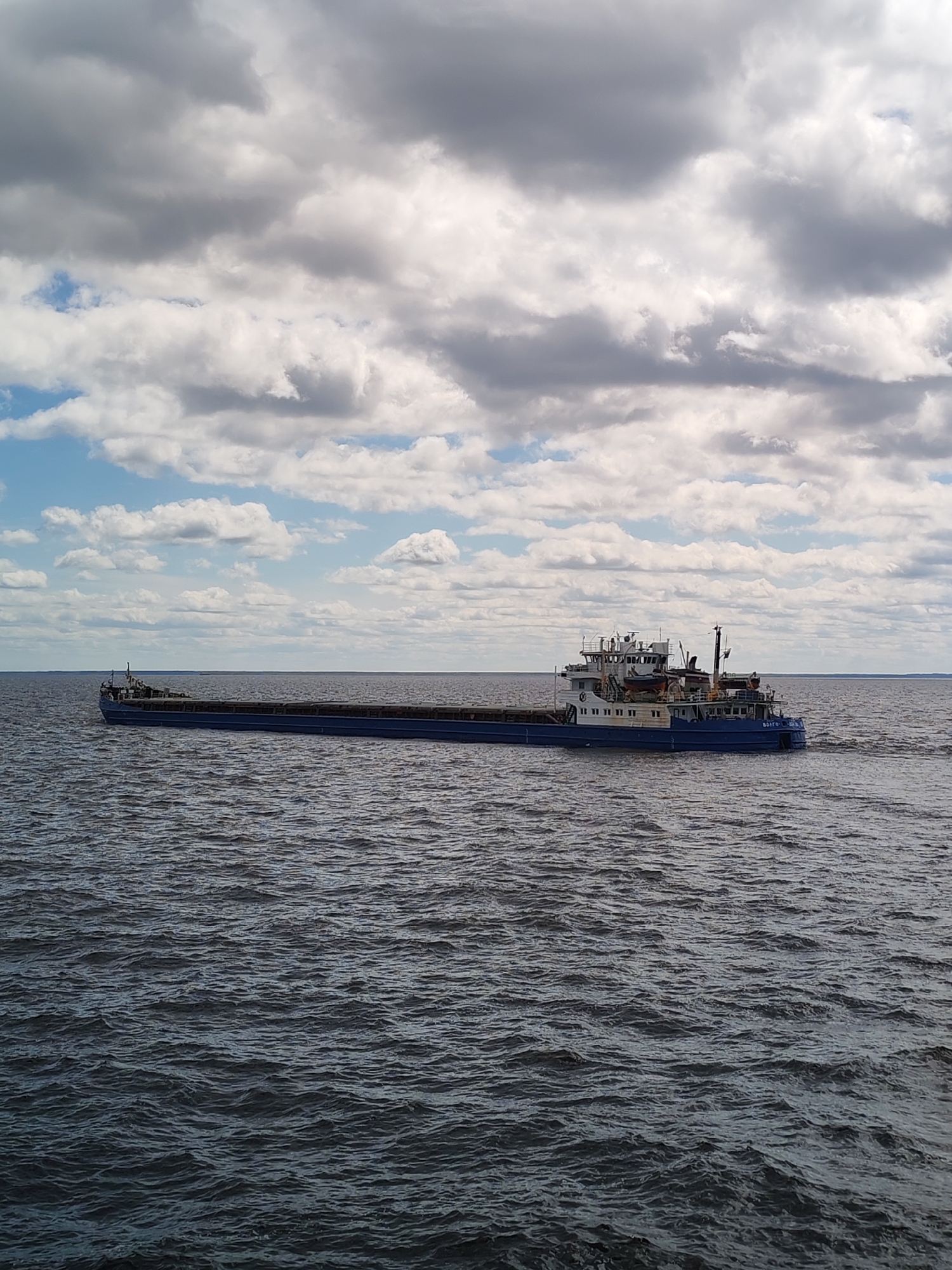 A wide shot shows a long blue and white barge moving on water. The barge has a control cabin on the right side with a visible name in Cyrillic script, suggesting the location may be Russia. The water is dark and choppy. Above, the sky is filled with white and gray clouds with patches of blue sky peeking through. There are no people visible in the image. The photograph appears to be taken during the daytime under overcast conditions. No specific details indicating Mologa, Russia are present within the imagery.