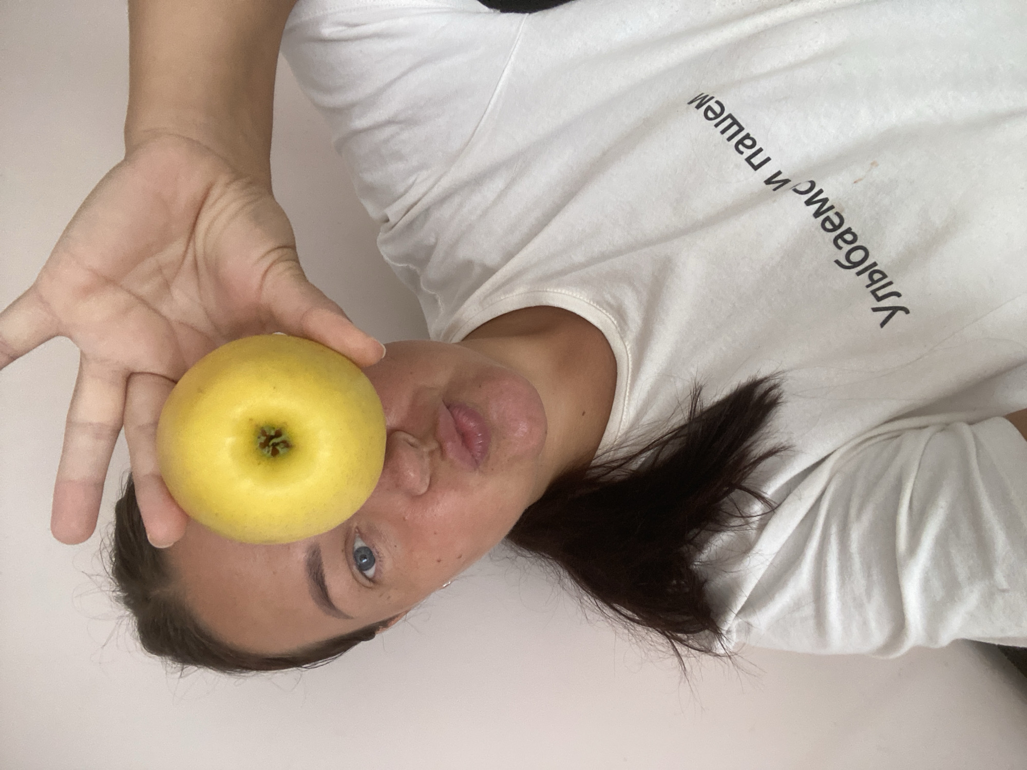 A woman with dark hair pulled back in a ponytail holds a yellow apple in front of her right eye, obscuring it. She is looking towards the camera with her lips pursed. She is wearing a white t-shirt with black text that appears to be in Cyrillic. The background is a plain, light-colored wall. There are no recognizable landmarks or indicators to suggest the location is the city of Sochi, Russia.