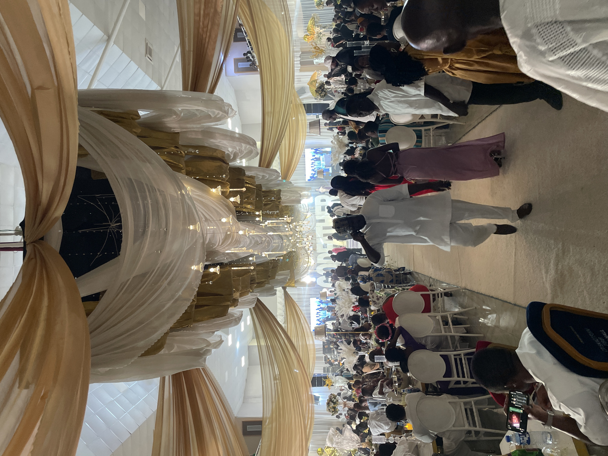 The image captures a large, indoor event in what appears to be Bar Jirgi Summa, Nigeria. Rows of seated people are arranged on either side of a central aisle. Most are dressed in white clothing, possibly for a ceremony or celebration. There is a man in white suit is walking down the aisle. Tables are positioned in front of the seated guests. Elaborate draping in white and gold fabrics adorns the space above the central aisle. Lighting fixtures are suspended from the ceiling. Some attendees appear to be using mobile phones. A screen displays information further down the room.