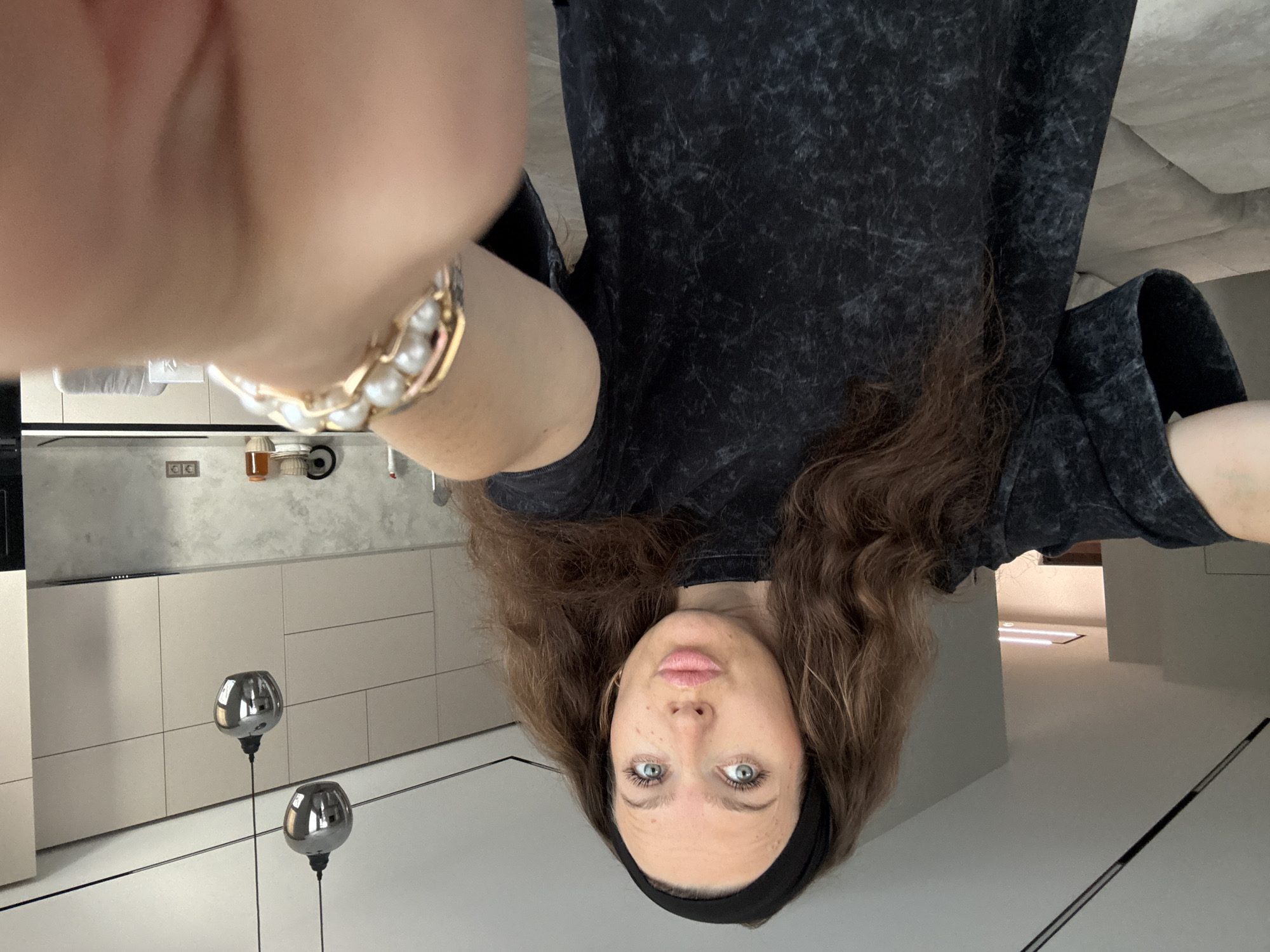 The image shows a woman taking a selfie in what appears to be a modern kitchen. The woman has long, wavy brown hair and is wearing a black t-shirt with a textured pattern. She is also wearing a black headband and a ring with pearls on her left hand. Her eyes are blue-green and she has a neutral expression.
The kitchen features sleek, light-colored cabinets and a grey countertop. Two spherical, metallic pendant lights hang in the foreground, reflecting the room. In the background, there are what look like kitchen appliances and a wall with electrical outlets. The overall aesthetic suggests a contemporary, minimalist design.
The lighting suggests it could be daytime, as there is ambient light coming from within the space and possibly from outside, although no windows are clearly visible. The scene is indoors, in a domestic setting. No text is visible in the image. The image is oriented vertically, with the woman's head at the bottom of the frame and her feet, if visible, would be at the top. The selfie is taken from a high angle, looking down at the woman.