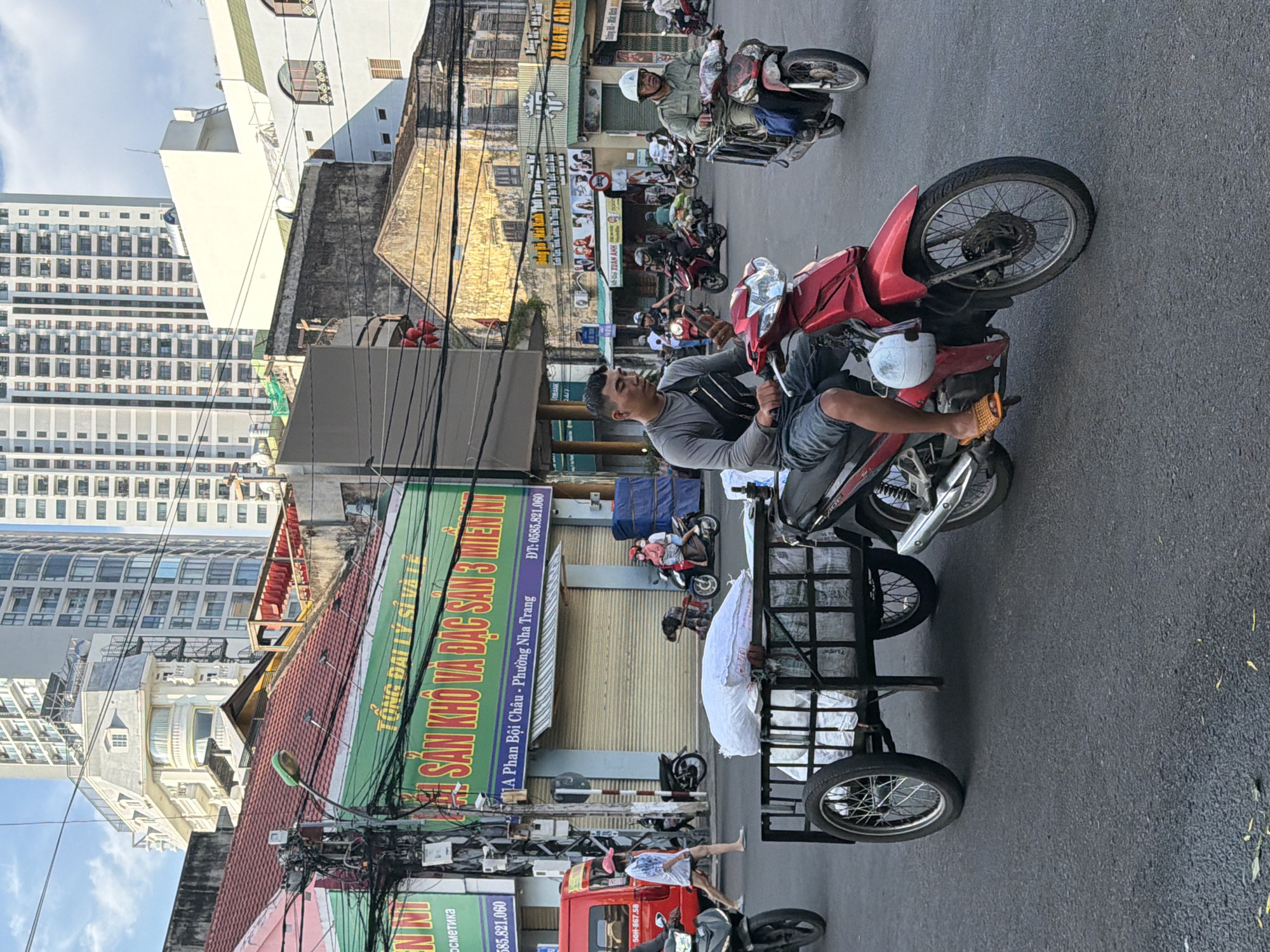 A street scene in Nha Trang, Vietnam, depicts urban traffic and commerce. In the foreground, a man on a red motorcycle pulls a trailer loaded with white sacks. He is looking at his phone, wearing a grey long-sleeved shirt, grey shorts, and orange sandals. Behind him, another motorcycle with a rider wearing a green shirt and helmet travels in the same direction. Further back, other motorcycles and a red van are visible on the street. The background features multi-story buildings, some with modern high-rise architecture and others with older, more traditional designs. Signs with Vietnamese text indicate businesses and commercial establishments. The sky is blue with scattered clouds. The overall impression is of a busy street environment in a Vietnamese city.