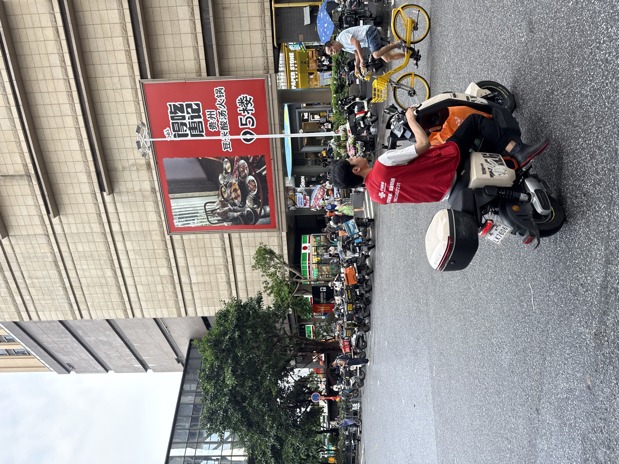 The image depicts a street scene in Haizhu District, China, with a focus on traffic and commercial activity. In the foreground, a person wearing a red vest with Chinese text and a logo is riding a scooter. The scooter has a beige body and a tan storage box on the back. The rider is wearing black pants and black shoes with red accents. To the right, another person is riding a bright yellow bicycle. This individual is wearing a light blue t-shirt and denim shorts. The background features a large building with a beige, tiled facade and a large red advertisement banner displaying food. Several parked scooters with delivery boxes are visible, suggesting a delivery or food service hub. Other pedestrians and vehicles are present, contributing to a bustling urban environment. The street is wet, indicating recent rain or a damp surface. The sky is overcast.
