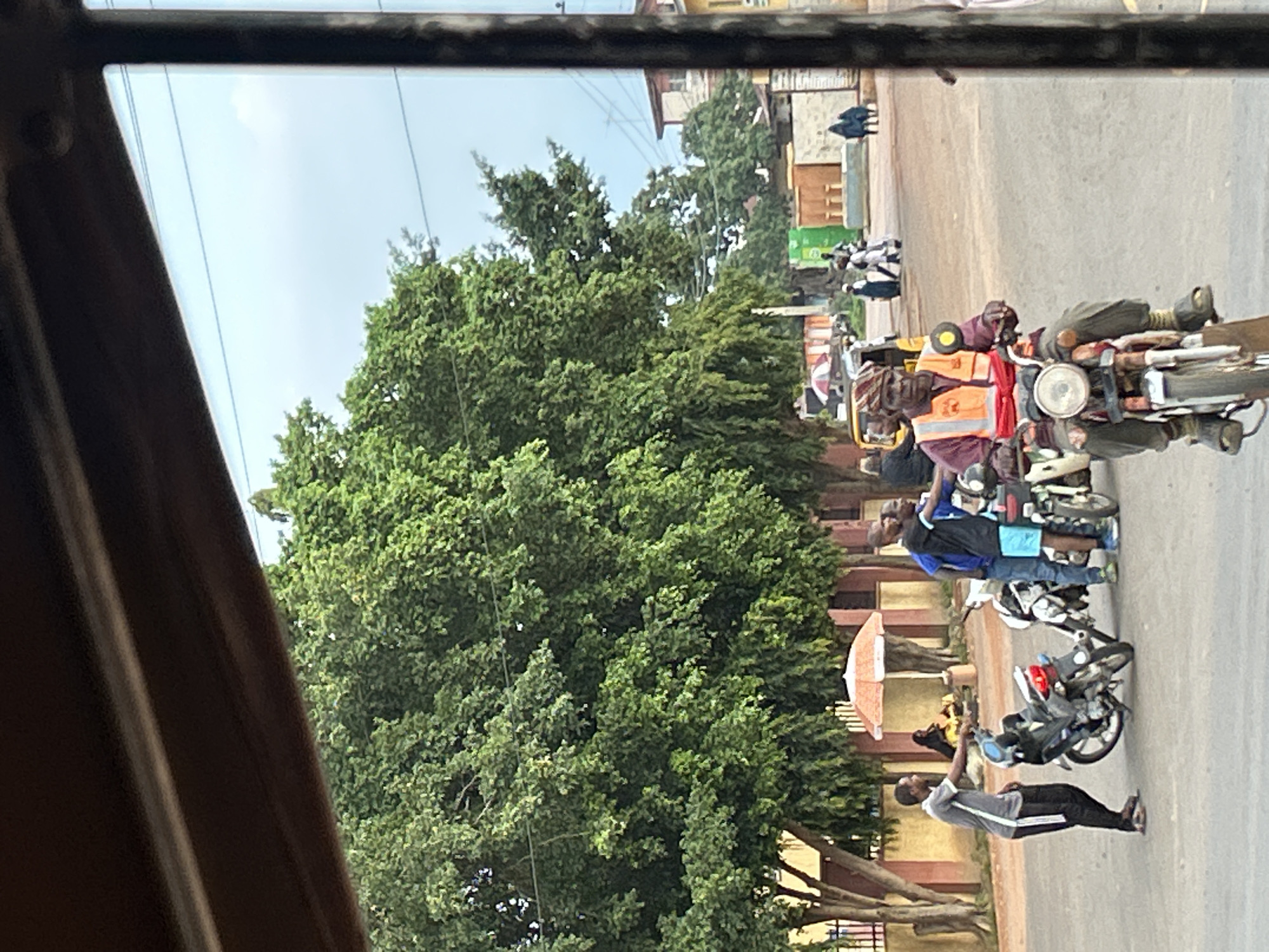This is a street scene in Kinkinau, Nigeria. The image is taken from inside a vehicle, looking out at a busy intersection. In the foreground, a man wearing an orange reflective vest and a patterned headscarf is riding a motorcycle. He is looking forward, with a neutral expression. The motorcycle is red and white. To the left of the motorcyclist, two men are standing next to a motorcycle. One man is wearing a grey t-shirt and black pants, while the other is wearing a blue t-shirt and blue shorts. Behind them, several other people are visible, including a woman sitting under an umbrella, and a group of people walking in the distance. The background is filled with lush green trees and buildings. The sky is overcast. The overall impression is of a typical day in a Nigerian city, with people going about their daily activities.