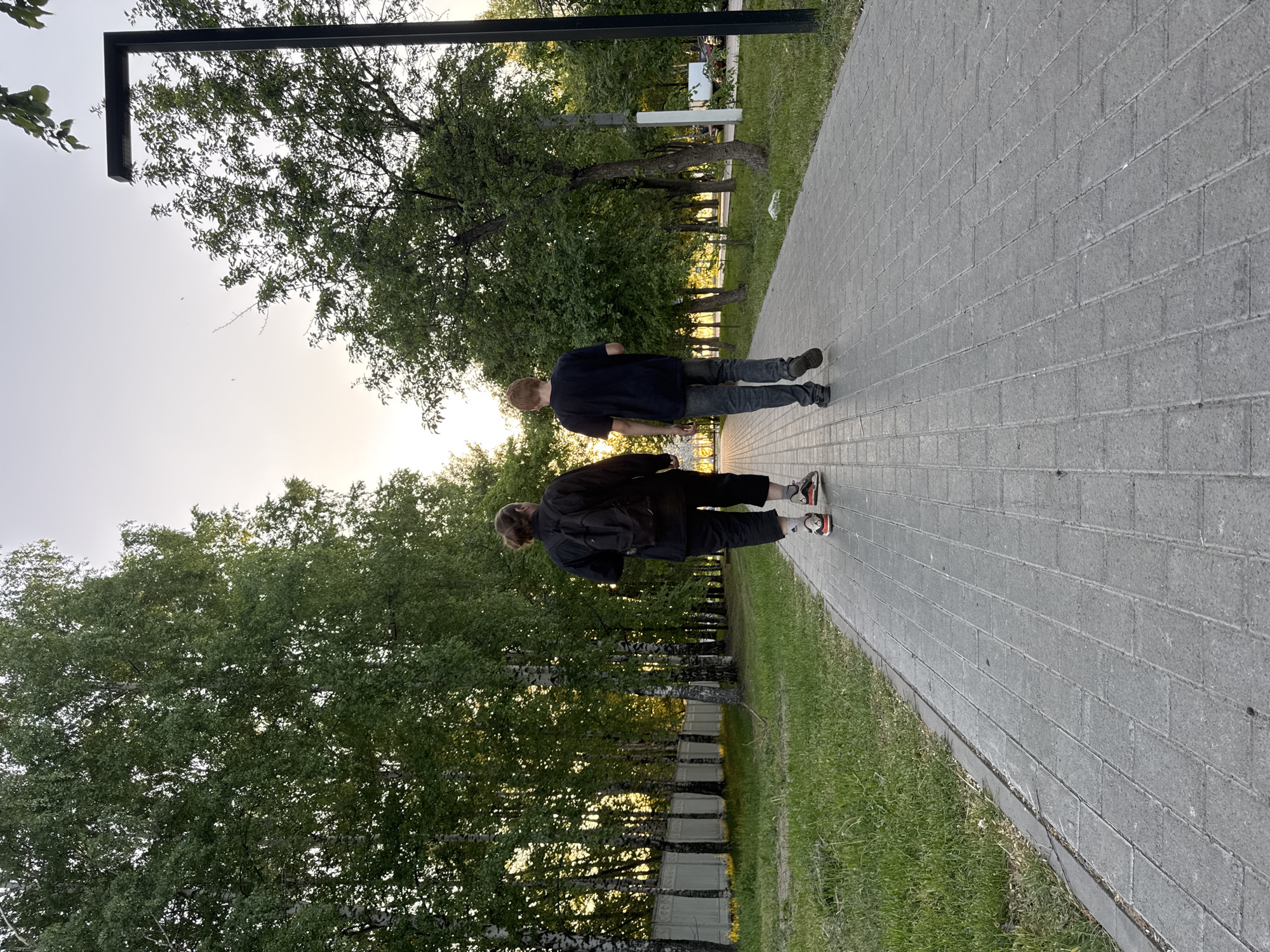 Two people are walking on a paved path through a park. The path is made of gray rectangular blocks. The person in the foreground is wearing dark clothing and sneakers with red and black accents. The person in the background is wearing a blue shirt and jeans. Trees line the path on both sides. The sun is setting in the distance, casting a warm glow on the scene. The location is in the city of Chaikovsky, Russia.