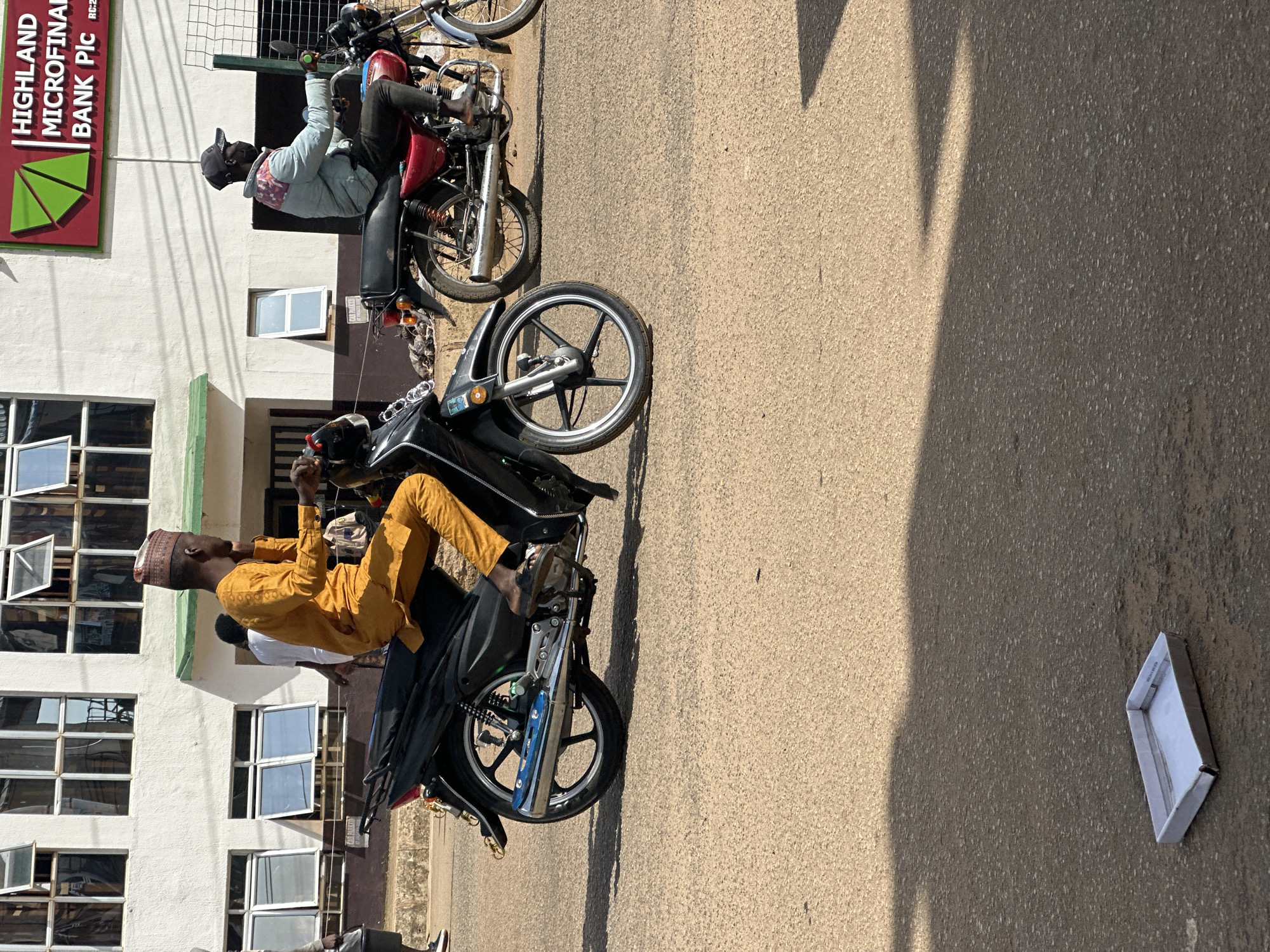In Bukuru, Nigeria, a street scene unfolds under bright sunlight. Two motorcycles are present. The one in the foreground, a black scooter, is ridden by a man wearing a yellow traditional outfit and a patterned cap. He is looking to his right. Behind him, another black motorcycle is visible, partially obscured. To the right, a red motorcycle is ridden by a man wearing a light blue jacket, dark pants, a black cap, and a face mask. He is looking forward.
In the background, a white building with multiple windows stands. A sign on the building reads "HIGHLAND MICROFINANCE BANK Plc". The street is paved with asphalt, and shadows are cast across it, indicating a sunny day. A discarded white tray lies on the asphalt in the foreground.