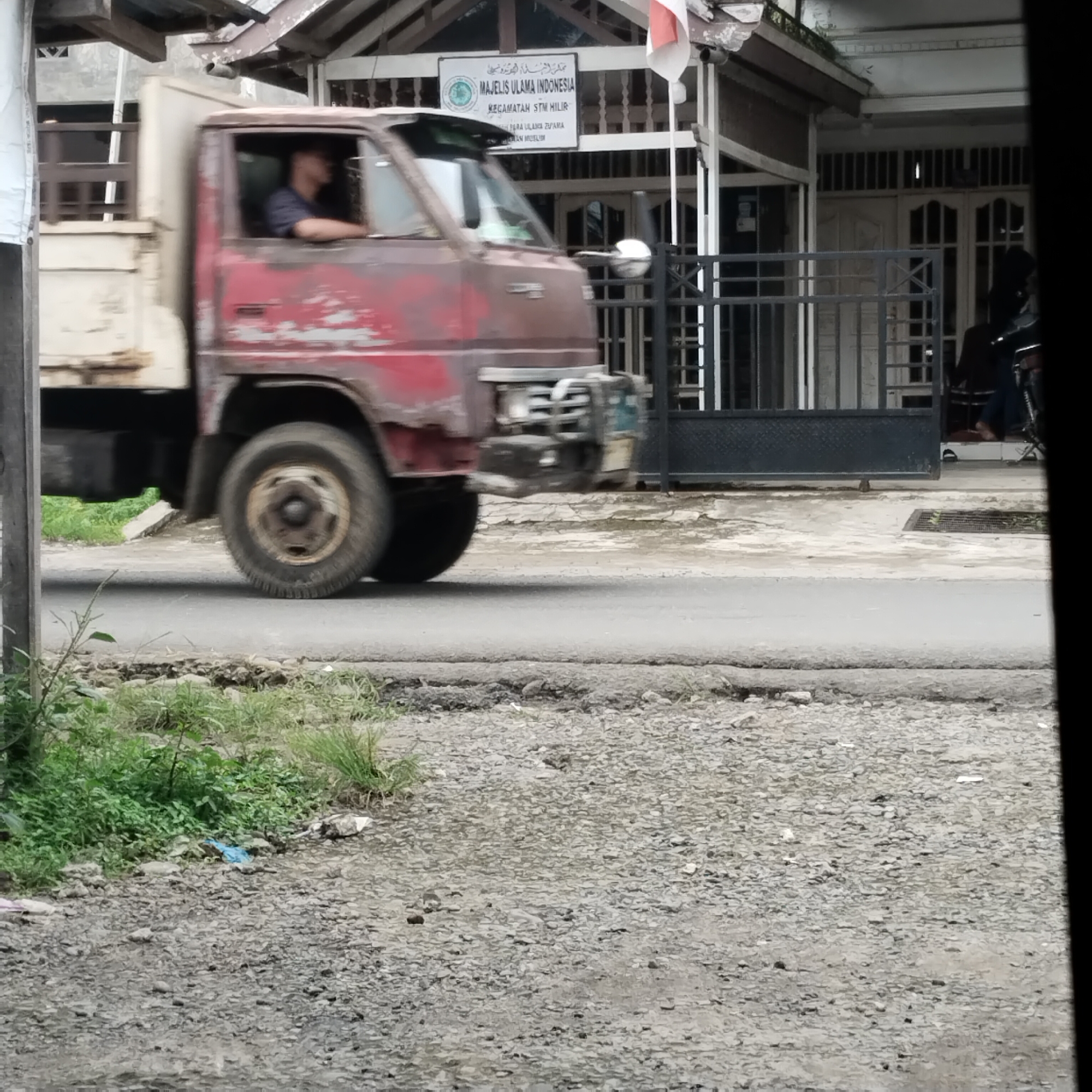 A faded red truck with a white cargo bed is moving from left to right in front of a building. The truck is the primary subject in the foreground. A person wearing a blue shirt is visible in the driver's seat. The building behind the truck has a sign that reads "MAJELIS ULAMA INDONESIA". There is a black metal gate in front of the building. The ground in front of the building and on the side of the road is covered in gravel and some vegetation. The road itself appears to be asphalt and has some cracks and patches. The overall impression is that of a street scene in a somewhat rural or less developed area of Talun Kenas, Indonesia.