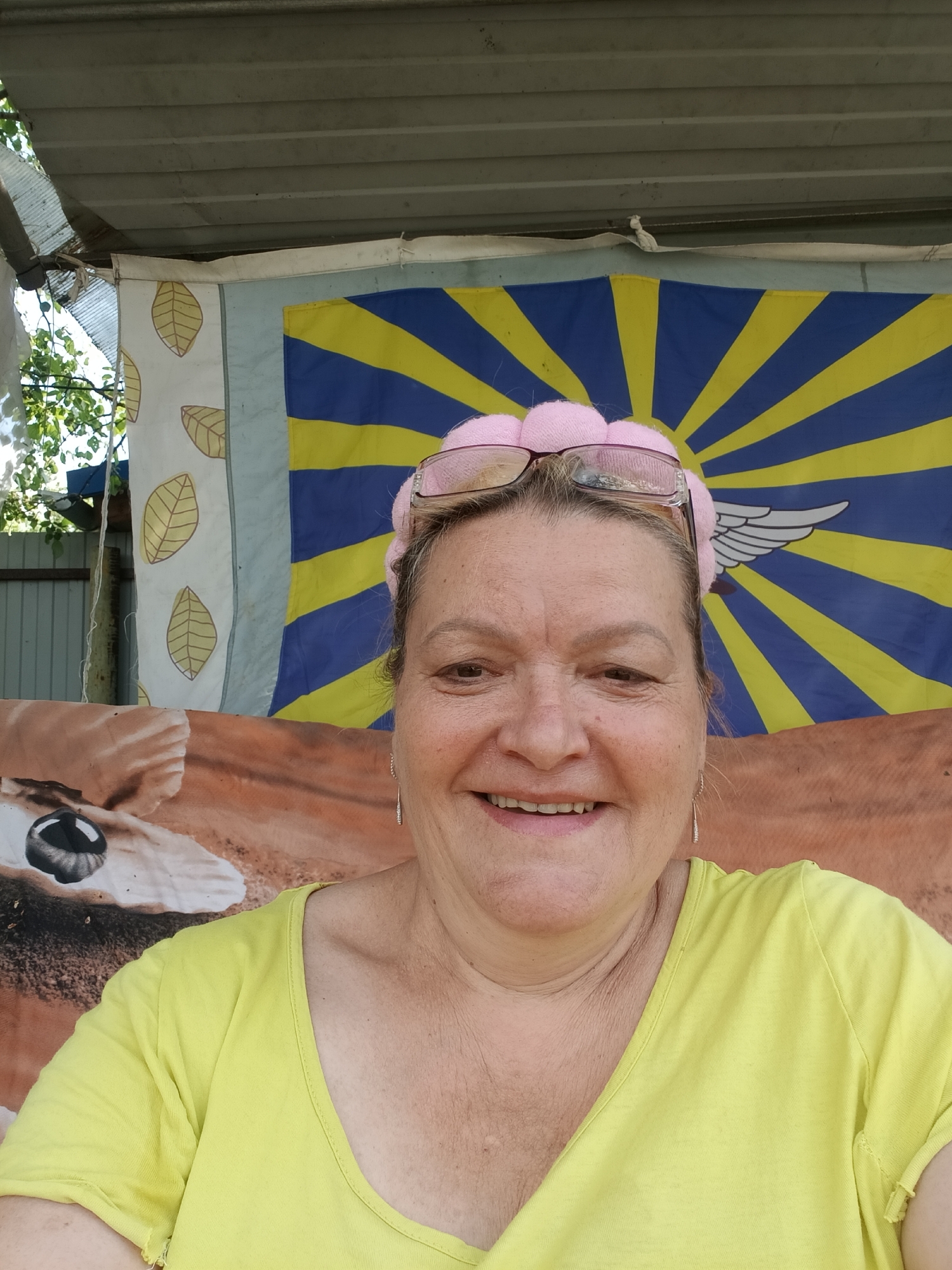 A medium shot depicts a smiling middle-aged woman with short brown hair, wearing a pink headband and glasses, and a yellow short-sleeved shirt. She is facing the camera. Behind her is a makeshift backdrop featuring various patterns. To her left is a white sheet with golden leaf patterns, and next to it is a blue and yellow sunburst design with a white wing emblem. Below these, there's a brown fabric displaying what appears to be an animal print with a prominent eye. The woman is likely standing under a corrugated metal roof, judging by the structure visible at the top of the frame. The image suggests an outdoor setting, potentially in Красный Маяк, Russia.
