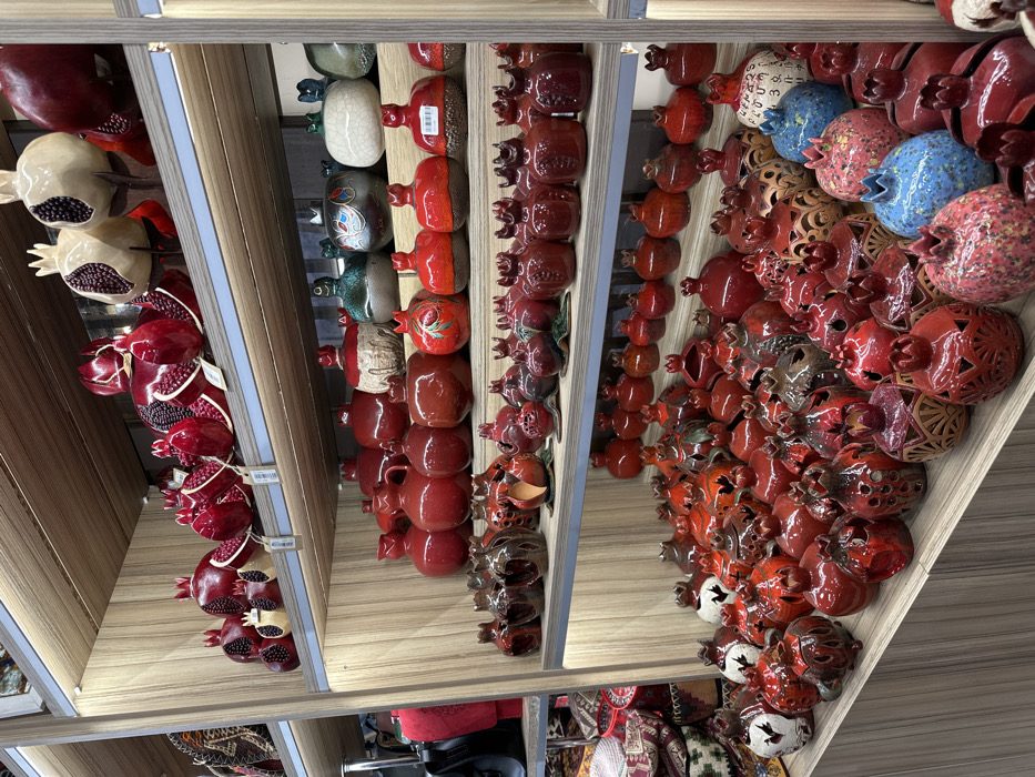 The image displays a detailed view of multiple wooden shelves laden with decorative ceramic and clay items, predominantly in the shape of pomegranates, located within an indoor retail space in Yerevan, Armenia. The shelves are light brown with vertical grain and are illuminated by integrated strip lighting positioned beneath each tier.
The top shelf features larger, glossy red and cream-colored pomegranate sculptures, some depicted as split open with visible seeds. The second shelf from the top holds an assortment of glossy red ceramic jugs and rounded pomegranate-shaped vessels with textured cream, dark teal, and patterned finishes. The third shelf contains numerous smaller, deep red, and reddish-brown pomegranate items, some with carved details or cutout patterns. The fourth and lowest visible shelves continue this dense display, showcasing a variety of pomegranate-shaped pieces in red, brown, speckled blue, and speckled pink/red, many featuring intricate cut-out designs suggestive of lanterns or candle holders. One cream-colored pomegranate on the bottom shelf includes visible Armenian script. A black handbag and a red bag are partially visible on a lower shelf to the far left. No individuals are present in the frame; the items are static, arranged for retail presentation.