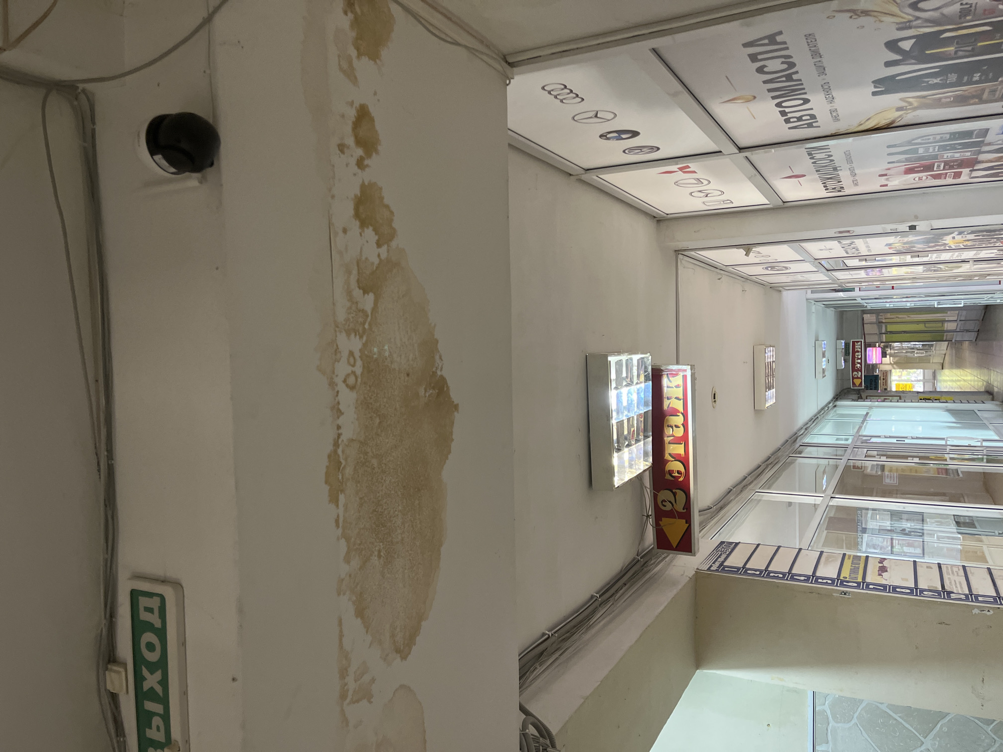 The image depicts an indoor corridor, likely within a commercial building. A prominent green exit sign is visible on the ceiling. A security camera is mounted on the ceiling, and there are signs of water damage on the ceiling surface, appearing as discolored patches. A red sign with yellow arrows and Russian text indicating "2 этаж" (2nd floor) is hanging from the ceiling. Along the walls of the corridor, there are storefronts or offices with glass doors and windows, displaying various advertisements, including logos of car manufacturers and promotional material for "АВТОМАСЛА" (motor oils) and "АВТОЖИДКОСТИ" (automotive fluids). A list with numbered entries, possibly a directory or price list, is attached to a column on the left. The overall environment suggests a commercial or public space, potentially a shopping arcade or office building. There are no people visible in the image. The specific location in Ivanovo, Russia, cannot be determined from the visual information provided.