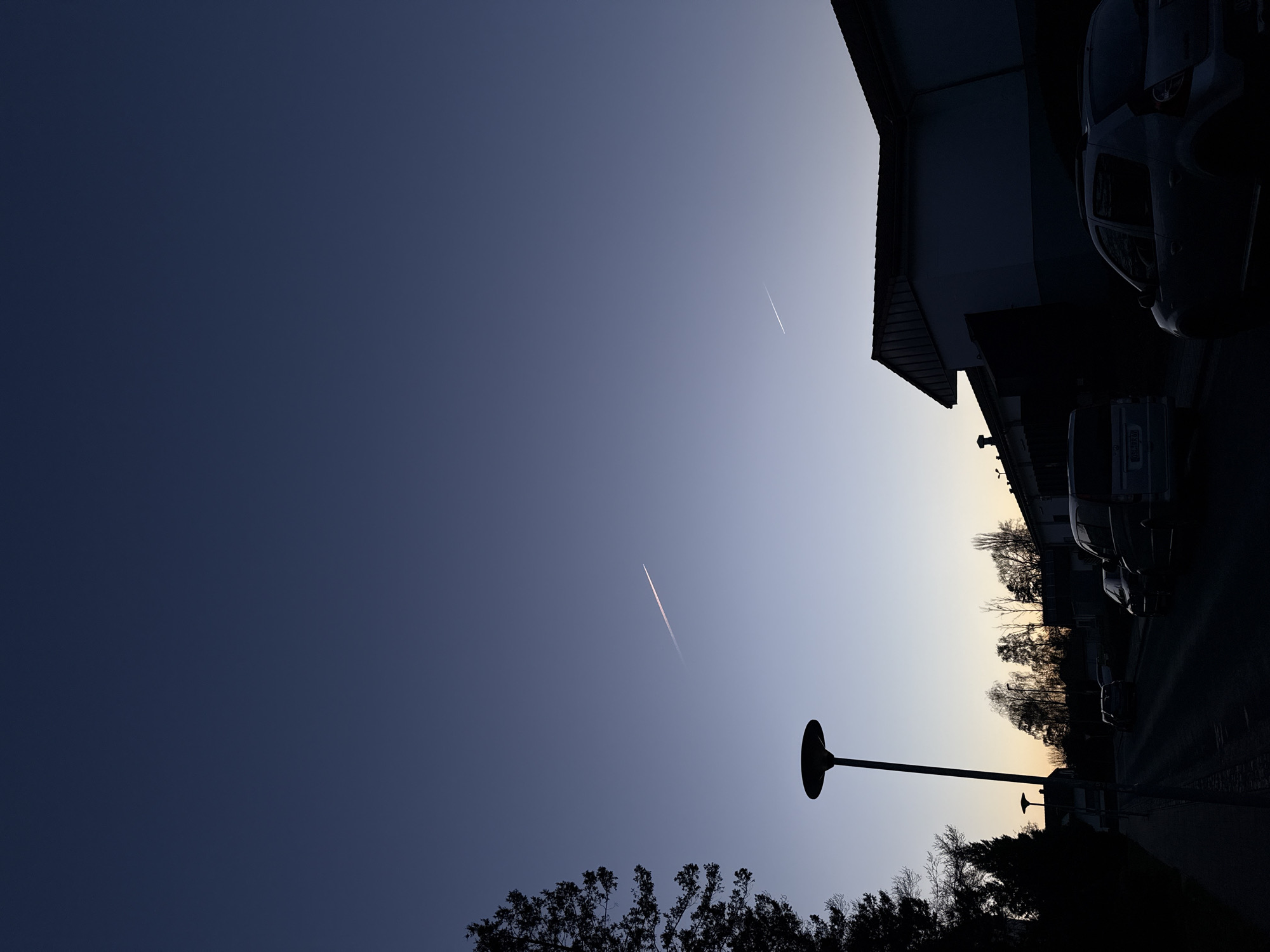 The image shows a street view in Lauchhammer, Germany, at twilight. The sky is a gradient from dark blue to light blue near the horizon. Two contrails from airplanes are visible in the sky. On the right side of the frame is a building and parked cars. A streetlight and trees are silhouetted in the foreground.