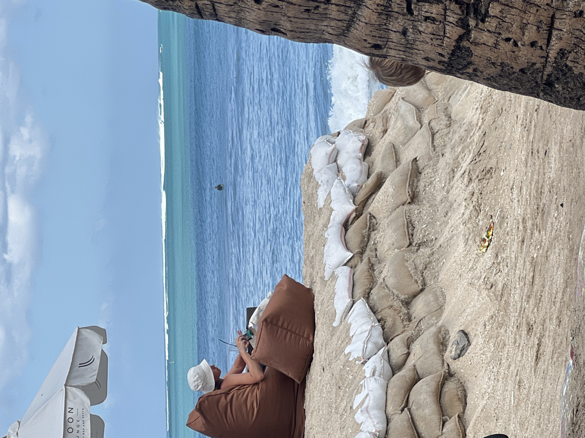 A person wearing a white bucket hat and a dark top sits on a large brown beanbag chair on a sandy beach. They are holding a drink and looking down at something in their hands. Behind them, the turquoise ocean stretches out to the horizon, where white waves are breaking. In the foreground, a wall of sandbags is stacked on the beach, with a few white bags on top. A large palm tree trunk is visible on the right side of the image, partially obscuring a blonde-haired child's head. The sky is blue with scattered white clouds. The scene suggests a relaxed day at the beach, possibly at a resort or cafe given the umbrella and beanbag chair. The location is Kuta, Indonesia.