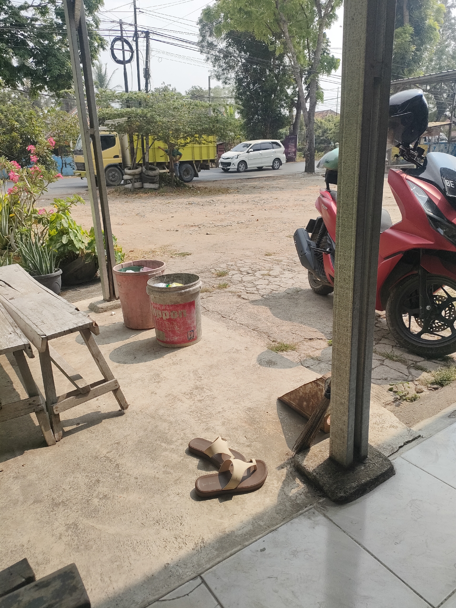 This image depicts an outdoor scene, likely in a residential or commercial area of Pringsewu, Indonesia. In the foreground, a weathered wooden bench is positioned on a concrete surface. To the right of the bench are two large plastic bins, one pink and the other a reddish-pink color, both appearing to be used for waste disposal. A red scooter with a black helmet secured to the back is parked on the right side of the frame, partially obscured by a concrete pillar. On the concrete ground near the pillar, a pair of light brown sandals are placed.
In the middle ground, a yellow truck is visible on a paved road, accompanied by a white van further down the road. Several trees with green foliage and some flowering plants with pink blossoms add to the natural environment. The ground surface in the middle ground appears to be a mix of dirt and cracked pavement. In the background, utility poles and overhead wires are prominent, indicating an urban or semi-urban setting. The overall lighting suggests it is daytime with bright sunlight casting shadows.