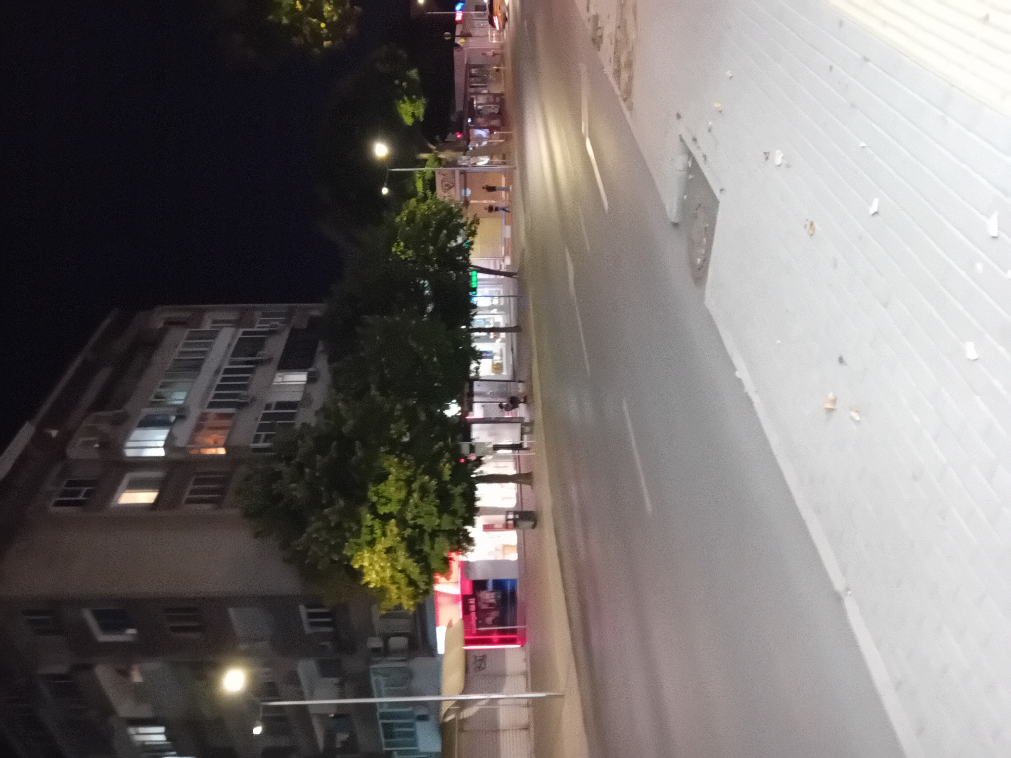 The image depicts a street scene at night in the city of Varna, Bulgaria. A multi-story apartment building with illuminated windows lines the left side of the street. Trees are planted along the sidewalk in front of the building. The street is paved and appears to be wet, reflecting the streetlights. To the right, there are shops with glowing signage, some of which are closed and shuttered. A bus stop shelter is visible, with a person sitting inside. Further down the street, there are more shops and streetlights. A yellow car is partially visible at the far right of the image, with its headlights on. The overall atmosphere is that of a quiet urban night, with artificial lighting illuminating the scene.