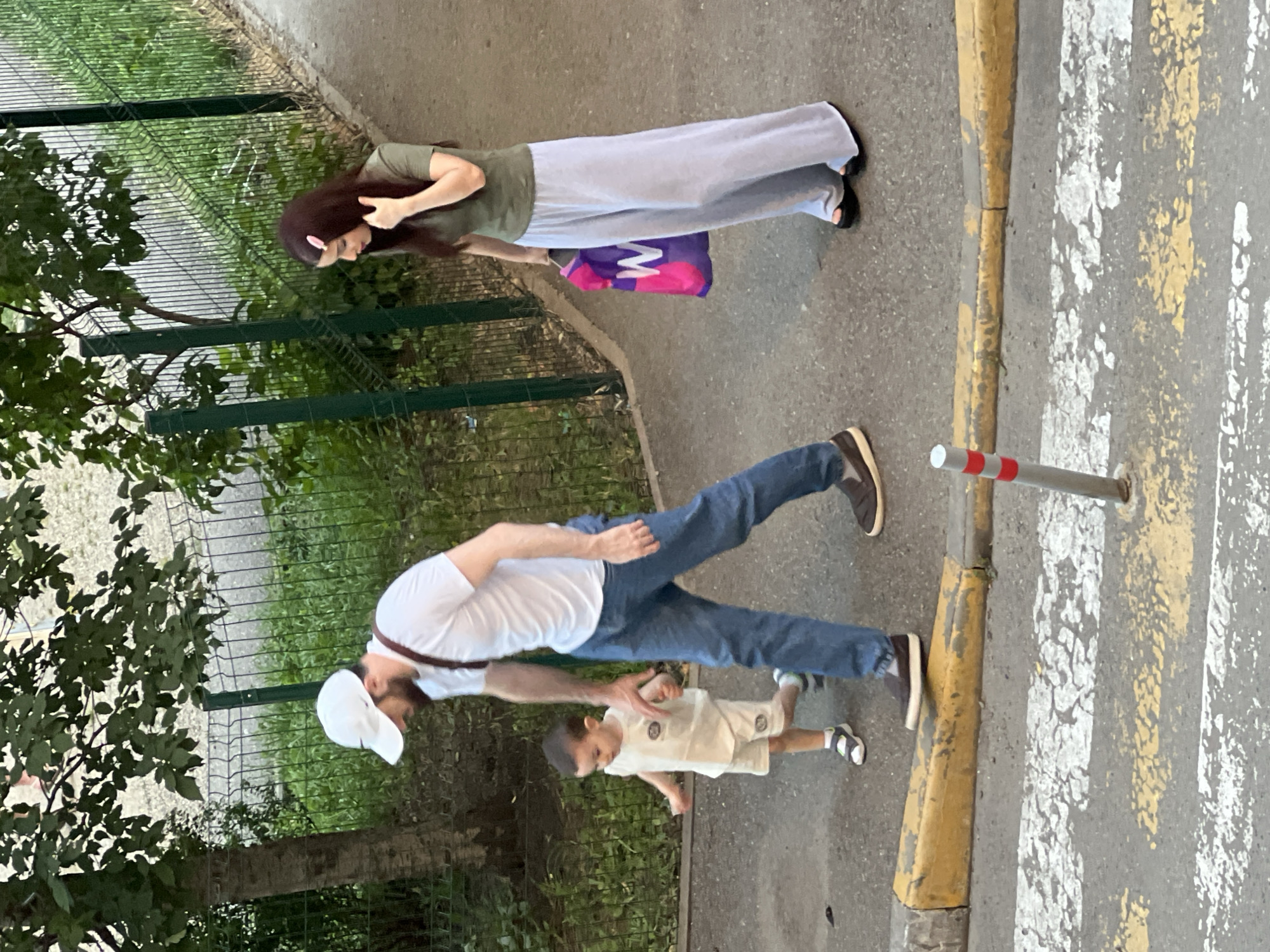 A man, a child, and a woman are walking on a sidewalk. The man, wearing a white t-shirt and jeans, is holding the child's hand. The child is wearing a white outfit and appears to be walking. The woman, in a green shirt and light blue pants, is carrying a pink and purple shopping bag with a white "W" on it. They are walking near a green metal fence and trees. The foreground shows a crosswalk with yellow and white markings and a yellow curb. The scene appears to be in a residential area of Krasnodar, Russia.