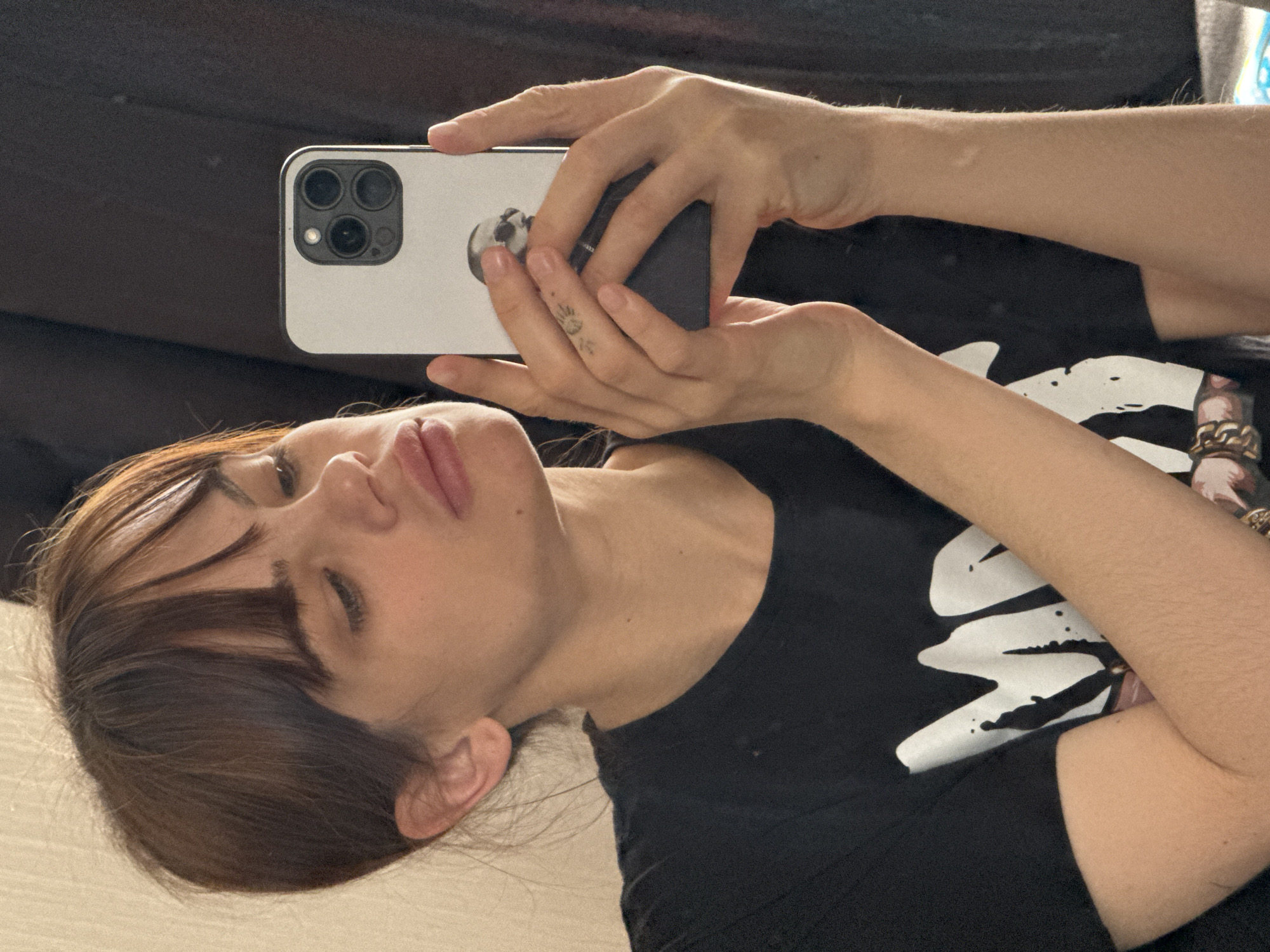 A woman with brown hair and bangs is lying down, taking a selfie with a white iPhone that has three camera lenses. She is wearing a black t-shirt. She has a small sun tattoo on her hand. The background appears to be a dark surface, possibly a couch or bed. The city of Rostov-on-Don, Russia is not discernible in the image.