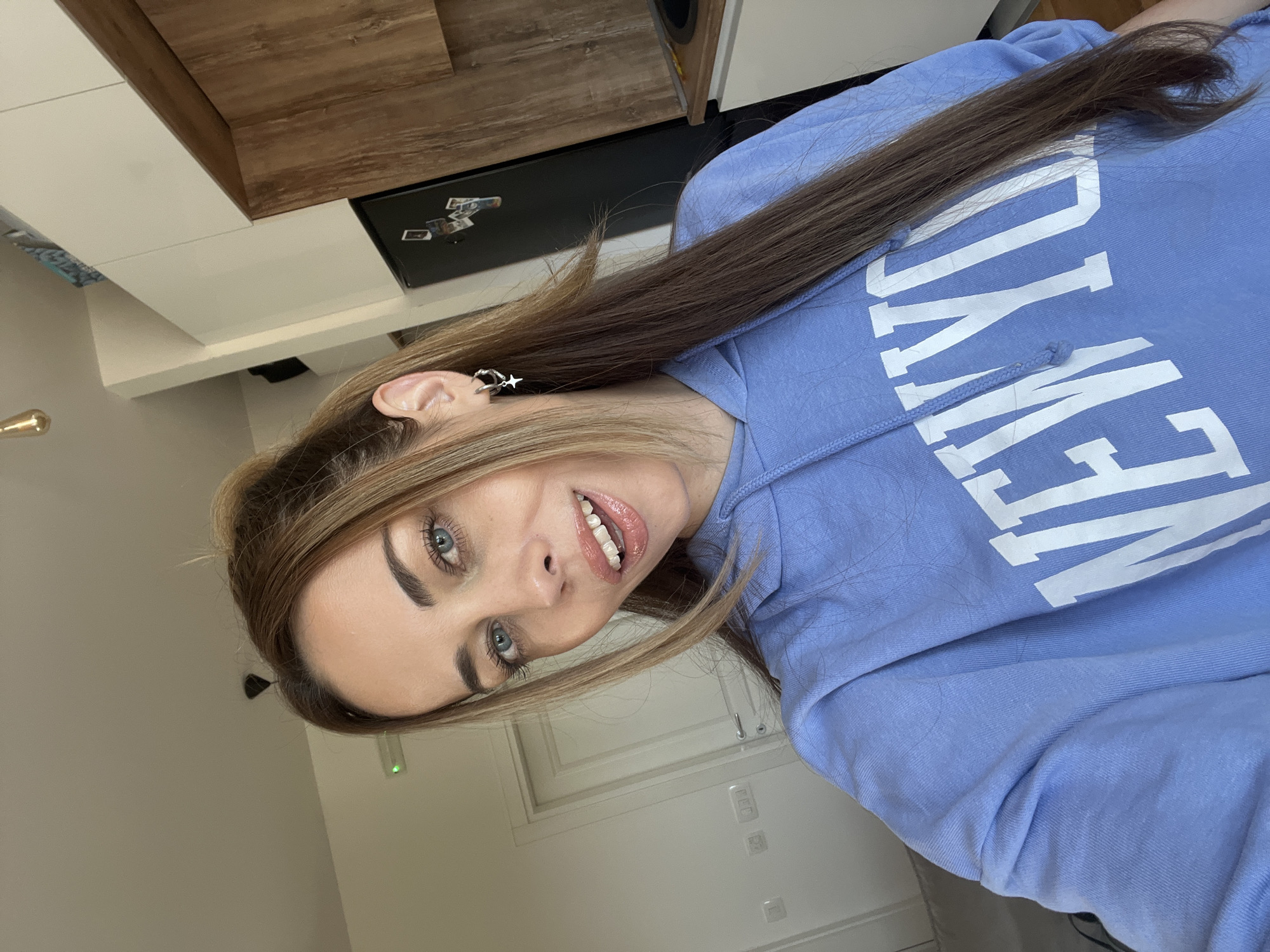 This image features a medium close-up of a young woman posing indoors, likely within a home setting in Bar, Montenegro. She looks directly at the camera with a cheerful, open-mouthed smile, revealing white teeth and glossy lips. Her striking blue eyes are prominent, framed by well-defined eyebrows.
She has long, straight, light brown hair with blonde highlights, parted on one side, cascading over her shoulders and chest. She is wearing a light blue hooded sweatshirt (hoodie) that has the word "NEW YORK" printed in large white letters across the front. A distinctive earring or ear cuff with a star-like charm is visible on her left ear.
The background suggests a modern kitchen or living area. White cabinetry or shelving is visible behind her, alongside a dark-colored appliance, possibly a refrigerator, which has small items (magnets or photos) attached to its surface. A brass-toned light fixture is partially visible on the left side of the frame. The indoor environment appears well-lit, though no specific time of day or external weather conditions are discernible. No specific cues within the image indicate its location as Bar, Montenegro, beyond the provided context.