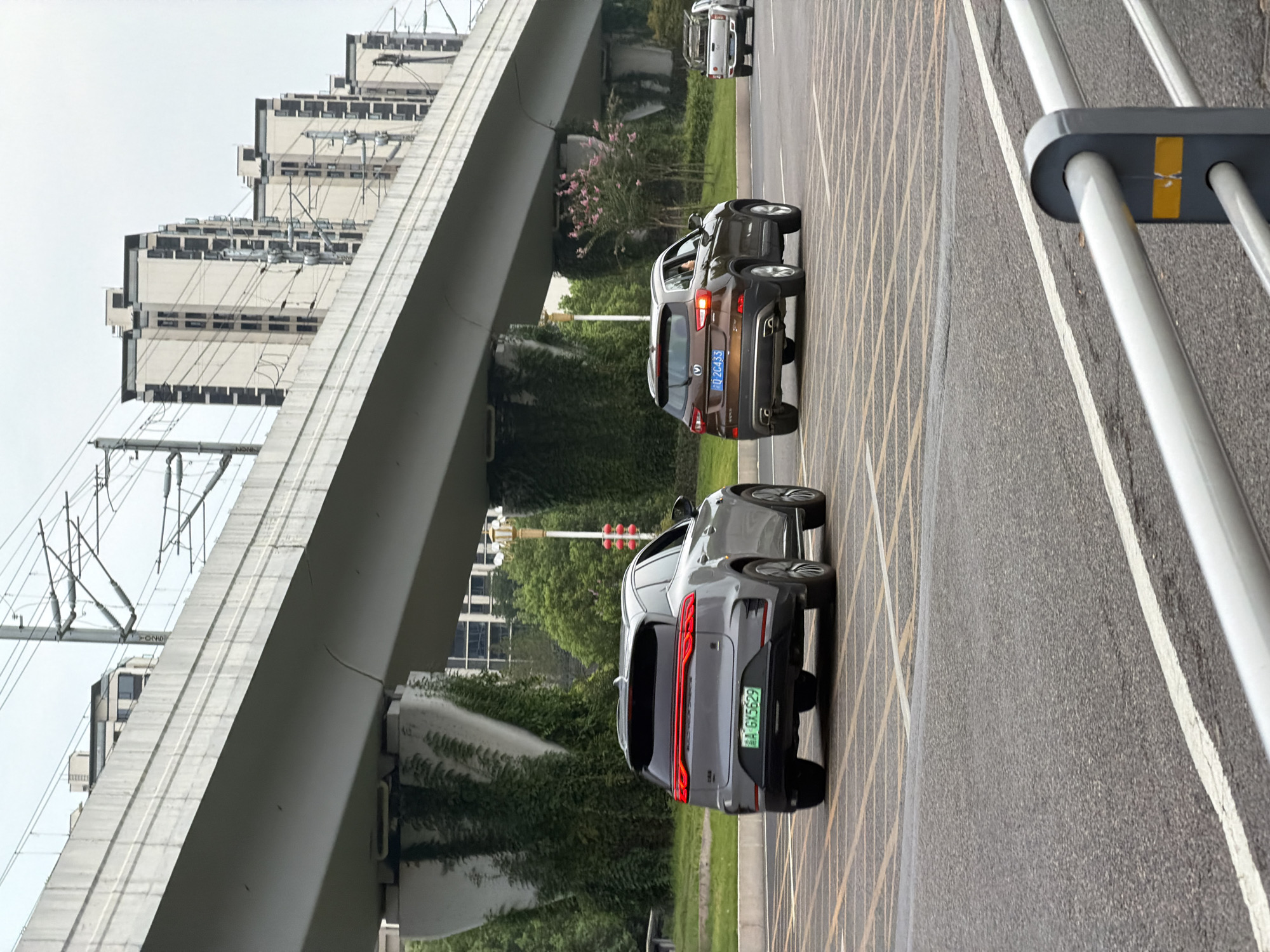 In Jiangjin, China, a grey SUV with license plate "AA GX5629" is traveling on a road. To its right, a brown SUV with license plate "2C433" is also on the road. In the background, an elevated concrete structure, likely part of an overpass or railway, spans across the scene. Buildings with a grid-like facade are visible behind the overpass, and trees and vegetation line the sides of the road. The sky is overcast.