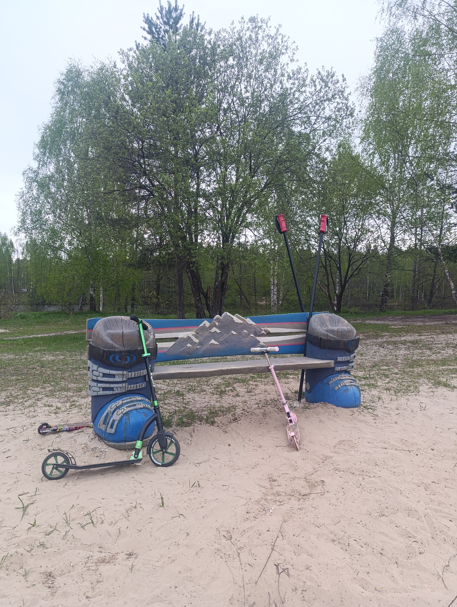 The image shows an outdoor scene in Chernogolovka, Russia. A park bench with a mountain scene painted on the backrest sits on a sandy area. The bench is flanked by two blue, stylized ski boot sculptures. A black and green scooter rests against the left side of the bench, and a pink scooter is on the right. A pair of black ski poles with red handles are propped up against the back of the bench. In the background, there is a grassy area bordered by trees. The sky is overcast.