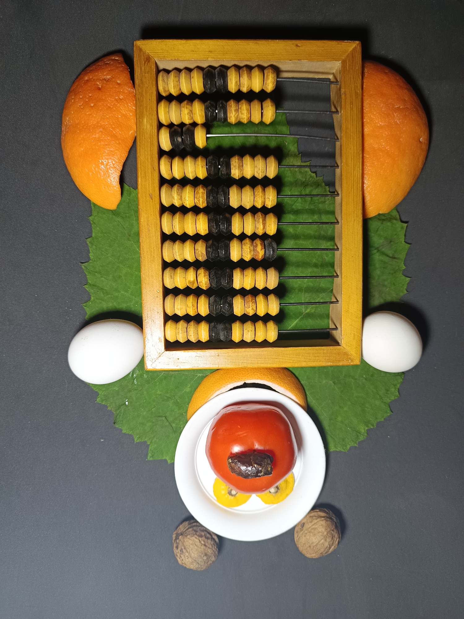 The image displays an arrangement of food items and an abacus on a dark gray surface, potentially representing a face or creature. Two orange slices are positioned on the left and right sides, serving as ears. Below them, two white eggs are placed, acting as cheeks. A large green leaf is situated centrally, beneath a wooden abacus with yellow and black beads. At the bottom, a white bowl contains a tomato, possibly representing the mouth, with two small yellow slices below it. Two walnuts are placed at the bottom corners of the image, resembling feet. The background is a plain, dark gray surface. The location in the city of Tyshky, Ukraine, is not determinable from the provided image. There are no people or actions depicted.