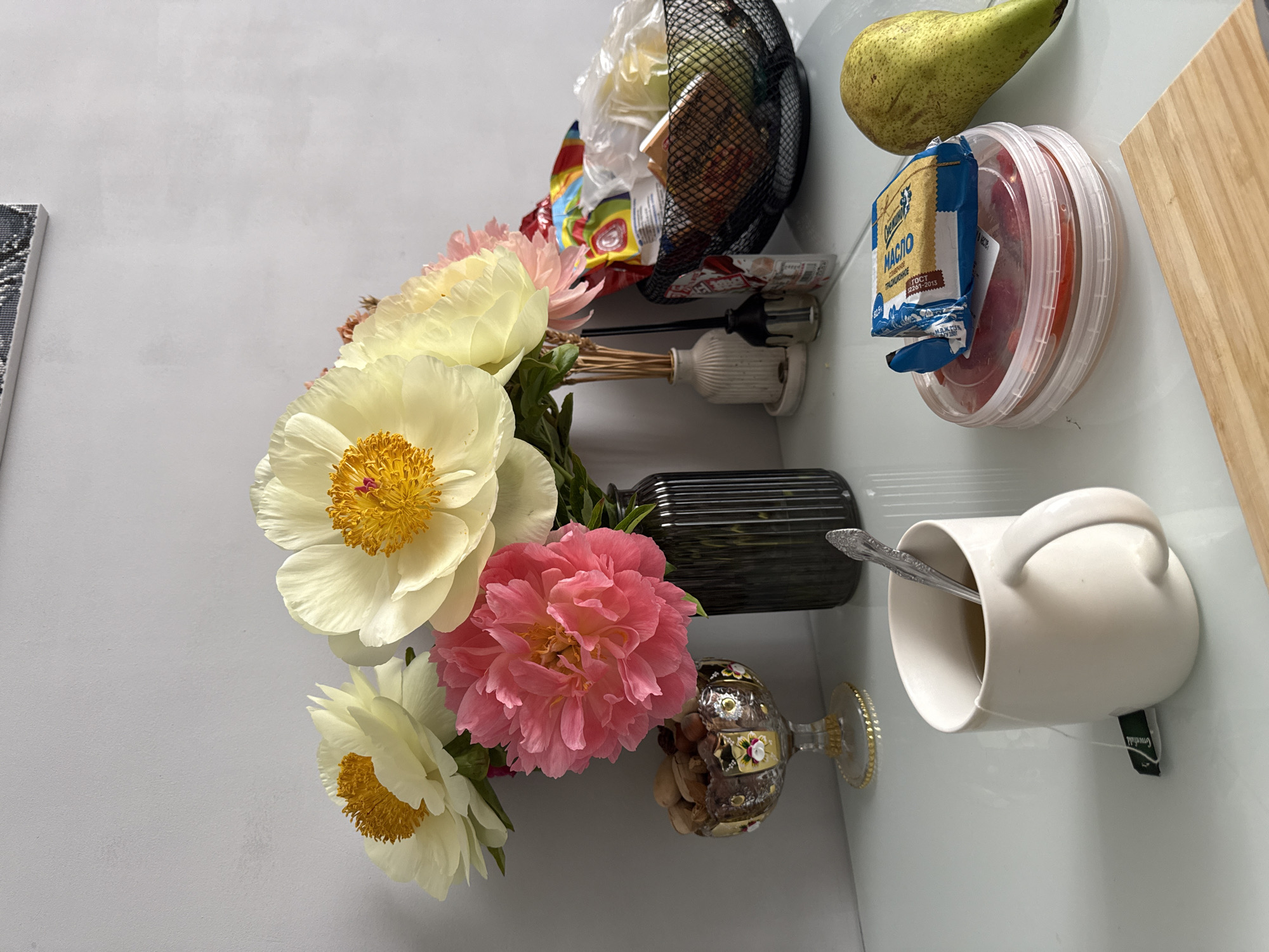 The scene depicts a still life arrangement on a white countertop. A bouquet of peonies, with both white and pink blossoms, is arranged in a collection of vases. A dark, ridged glass bottle serves as one vase, while another is a decorative, stemmed glass filled with nuts. Beside the flowers, a white mug with a spoon inside sits with a tea bag tag visible. To the right, a pear, a round plastic container with red contents (possibly tomatoes), and a package of butter are positioned. In the background, a black wire basket holds various items, including food packaging and what appears to be a lighter. A wooden cutting board is placed at the far right. The location is an interior setting in an unknown city.