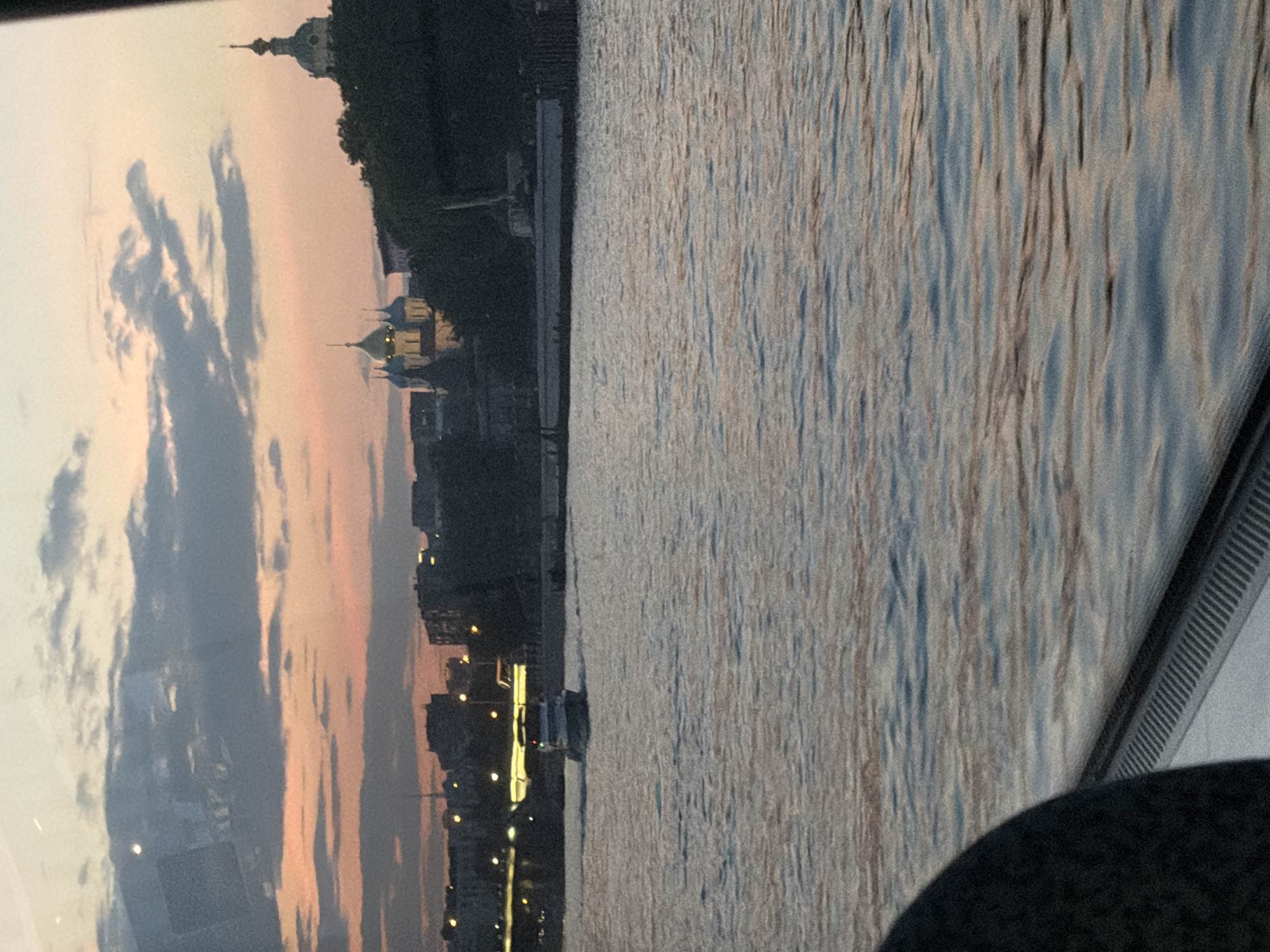 A boat is seen on the Moskva River at dusk, with the Moscow skyline in the background. The sky is filled with clouds, with hues of orange and pink from the setting sun. On the distant shore, buildings and the domes of churches can be seen. A bridge with lights is also visible in the distance. The water is calm with ripples reflecting the colors of the sky. A part of a vehicle's interior is in the foreground, obscuring the view to the left.