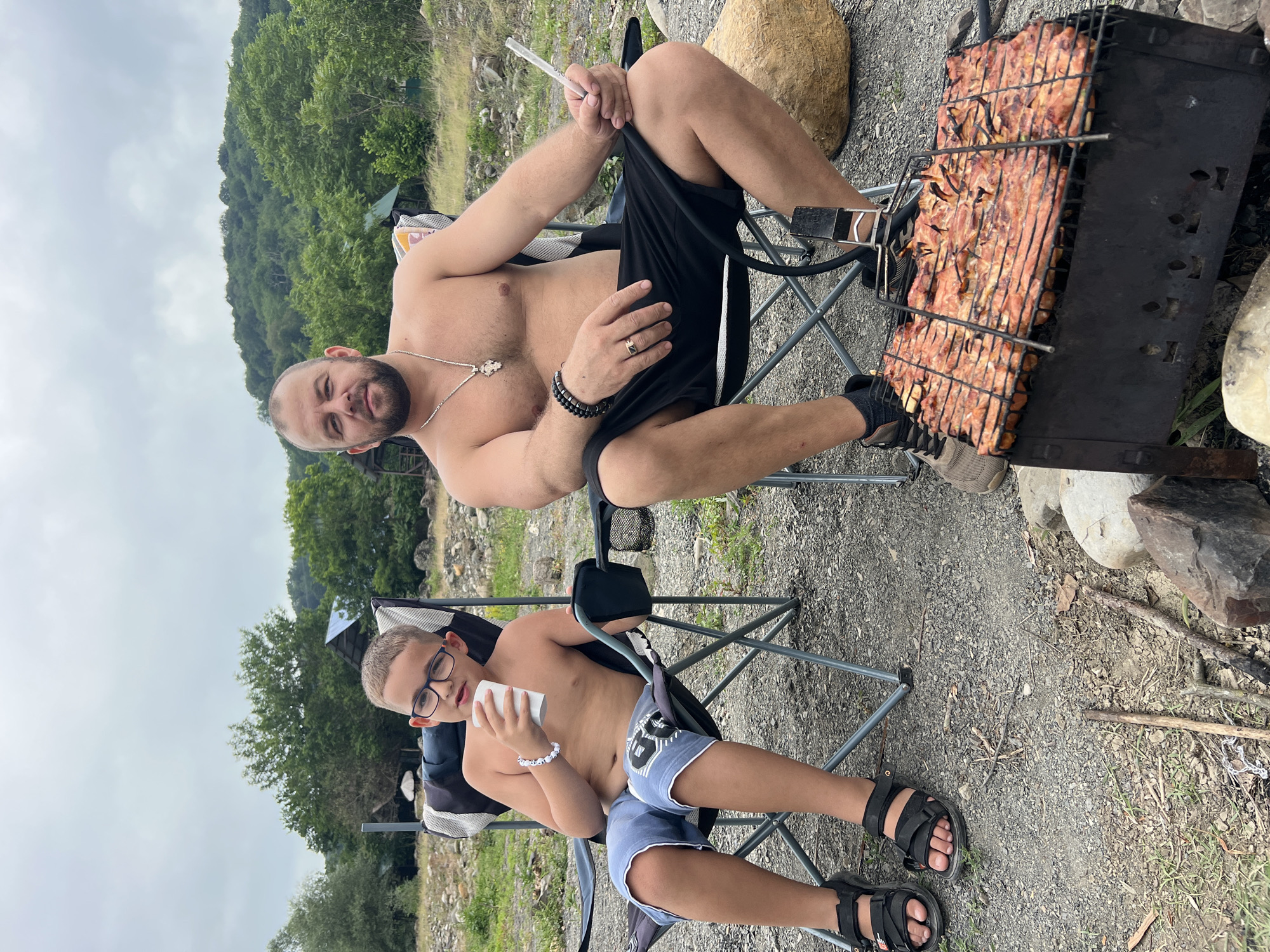 Two shirtless men, one an adult and the other a child, are sitting in camping chairs outdoors. The adult man is in the foreground, facing the camera with a beard and a necklace. He is holding a long, thin object in his right hand and wearing black shorts. Next to him, on the right, is a grill with meat cooking on it. The child is sitting in a chair in the lower part of the frame, wearing glasses and blue shorts. He is holding a white cup in his hands and wearing a beaded bracelet on his left wrist.
The scene appears to be a casual outdoor gathering, likely a picnic or barbecue, given the presence of the grill and the natural setting. The background shows a wooded hillside and a cloudy sky, suggesting it is daytime. The terrain is rocky and covered with gravel. The overall mood seems relaxed and informal. There is no visible text in the image. The location context is Sochi, Russia.
